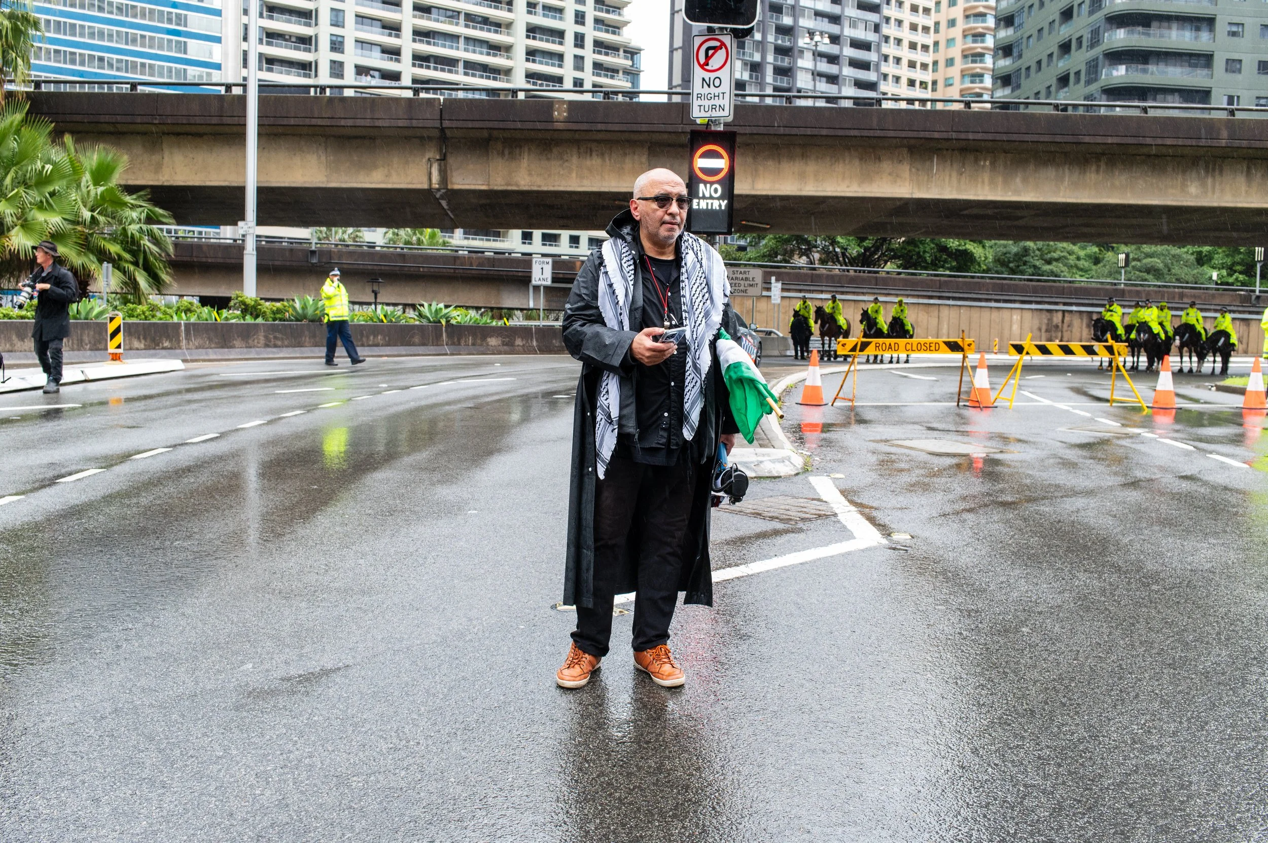 Man standing in front of a road block with police and mounted officers, wet pavement, city buildings in the background