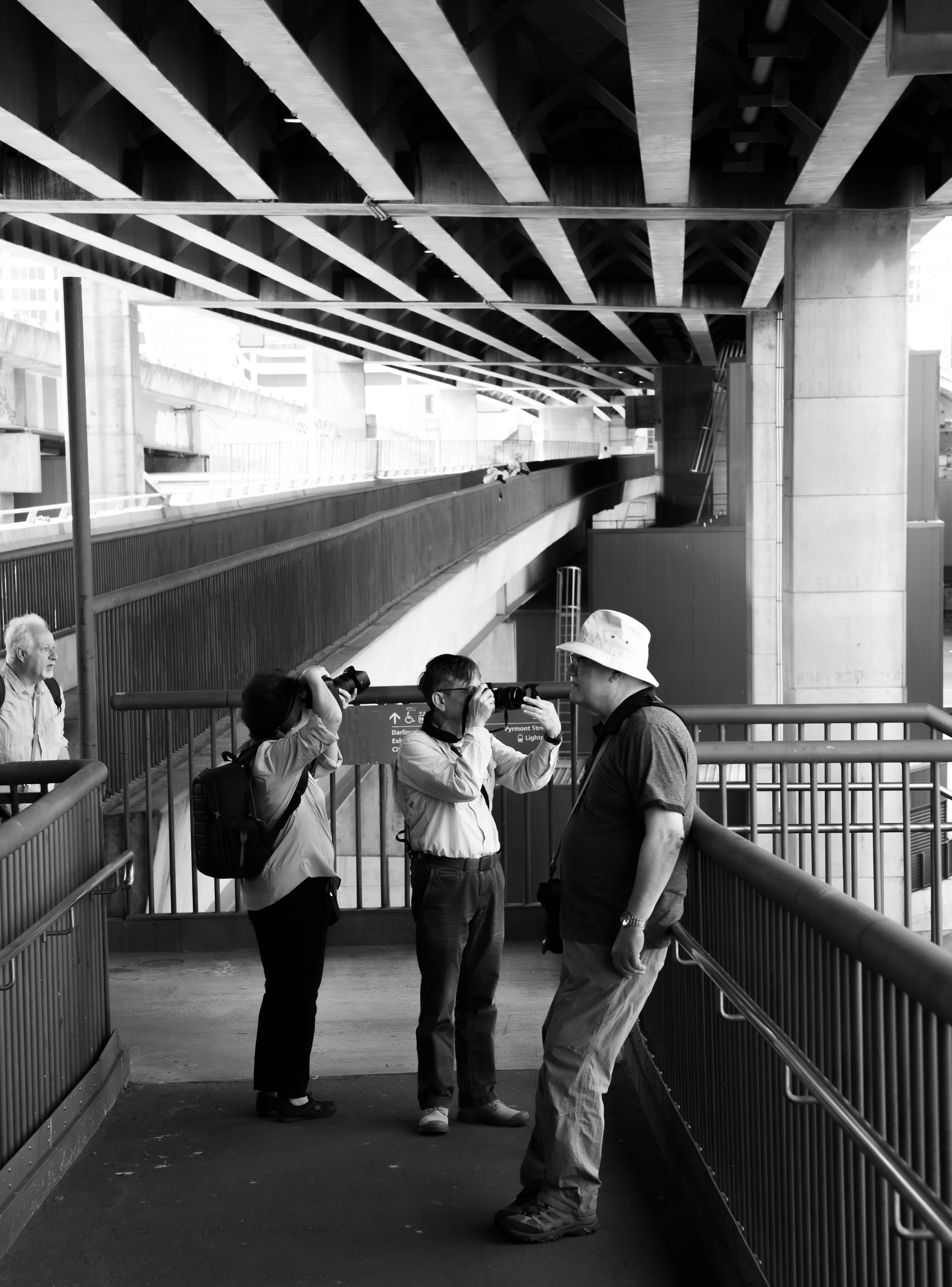 Three people standing on an outdoor walkway, two of whom are looking through binoculars, with one person facing them, all under a large overhead structure. Two other people are visible in the background.