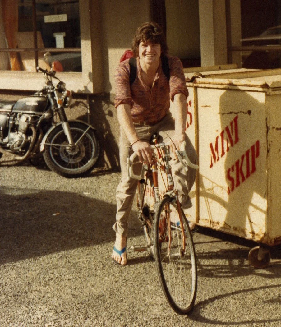 A young man with brown hair smiling, wearing a red patterned shirt, beige pants, and flip-flops, is pushing a bicycle on a gravel surface. Behind him is a black motorcycle and a yellow cart with red writing that says 'MONEY SKIP' near a building with a window.