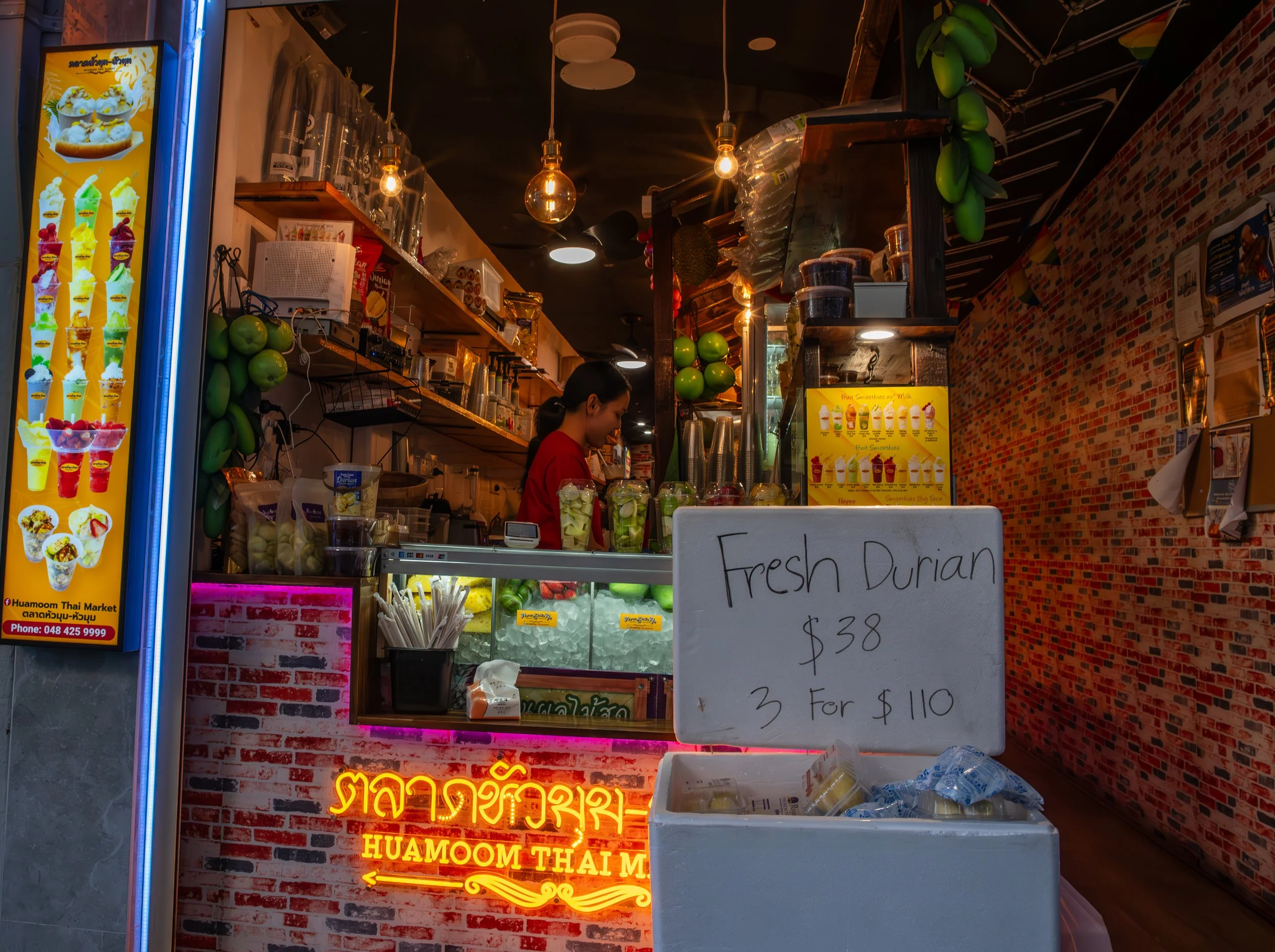 A Thai fruit stand selling fresh durian at $38 each or three for $110, with a woman working behind the counter surrounded by various containers and decorations, and a yellow menu display of fruit smoothies.