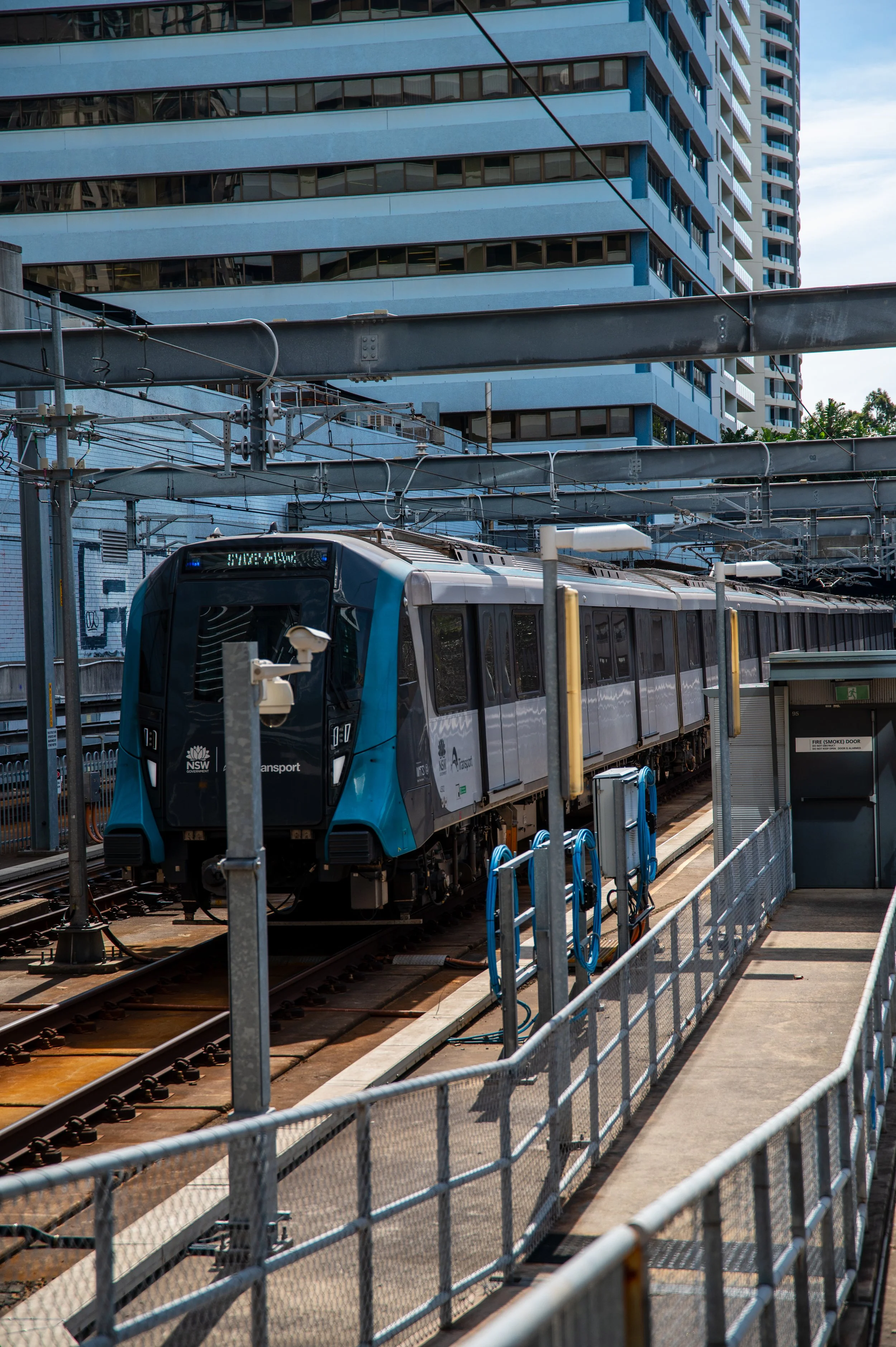 Modern blue and gray train on tracks at urban station with tall buildings and overhead wires in the background.