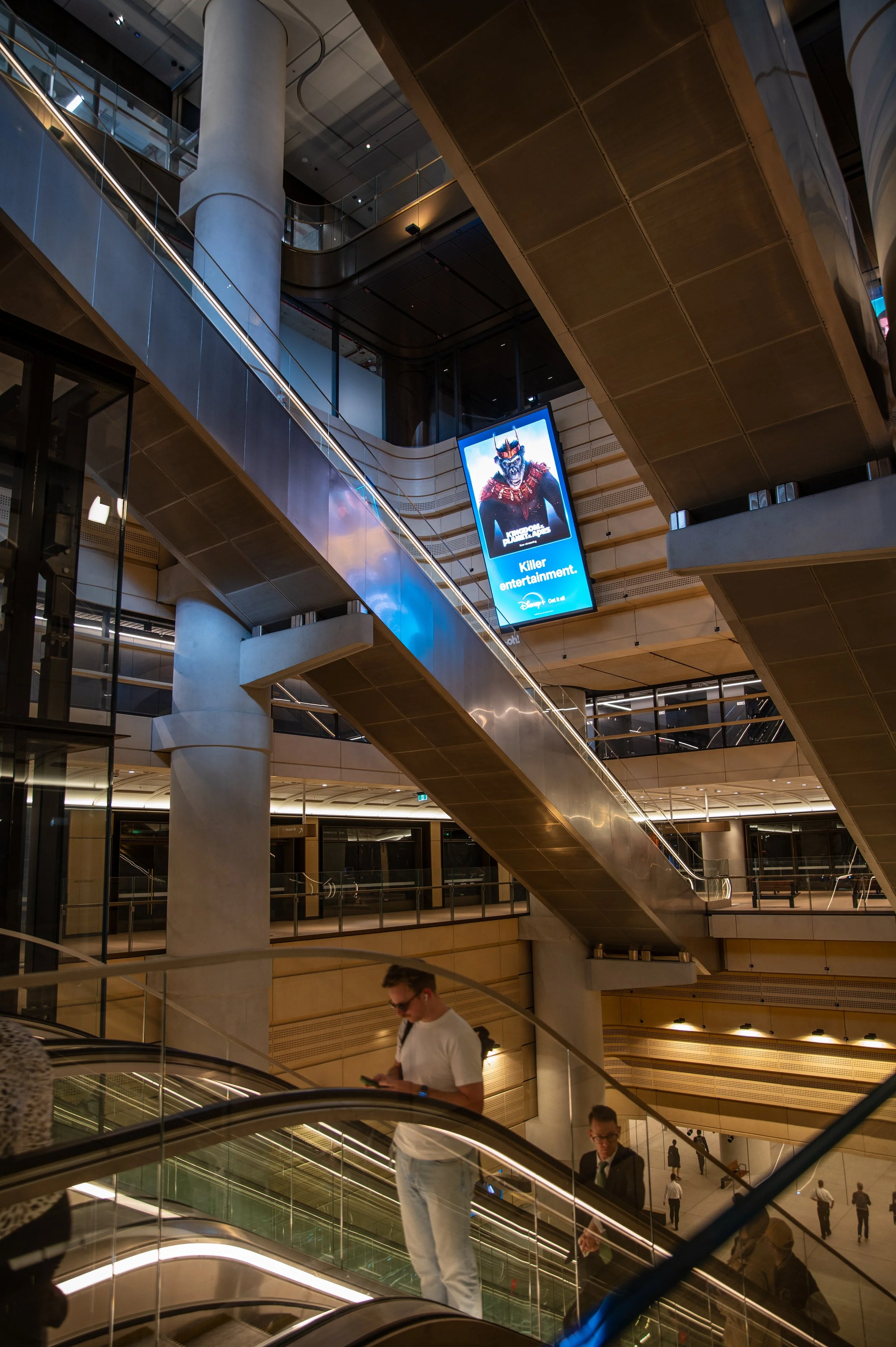 Interior view of a modern multi-level building with escalators, large digital screens, and people using their phones.