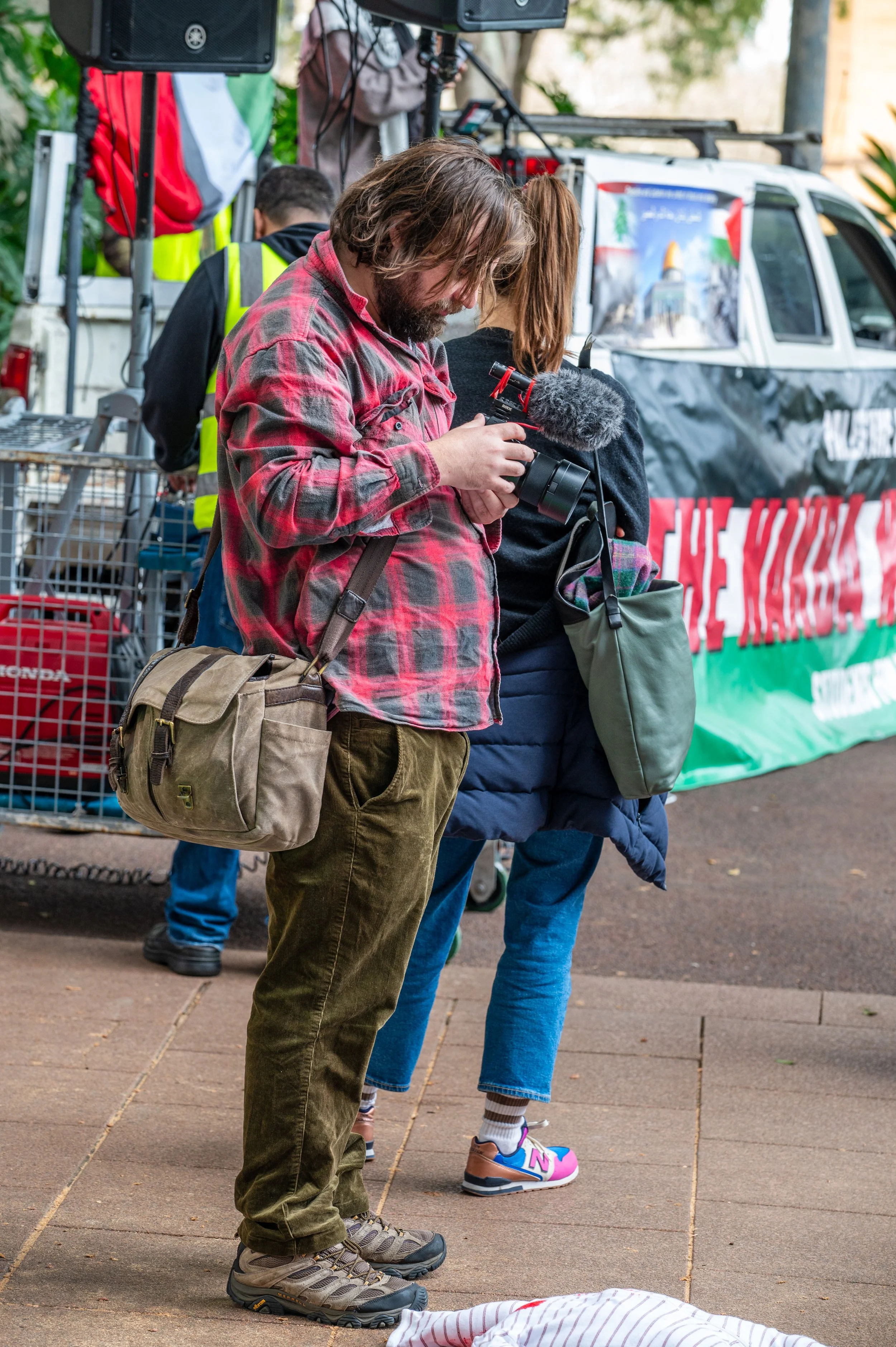 A man with a beard and long hair wearing a red plaid shirt and brown pants holding a professional camera with a microphone attached. He has a tan shoulder bag across his body. Behind him are people, some wearing yellow safety vests, near a vehicle with a banner that reads 'The March' and 'the', with trees in the background.