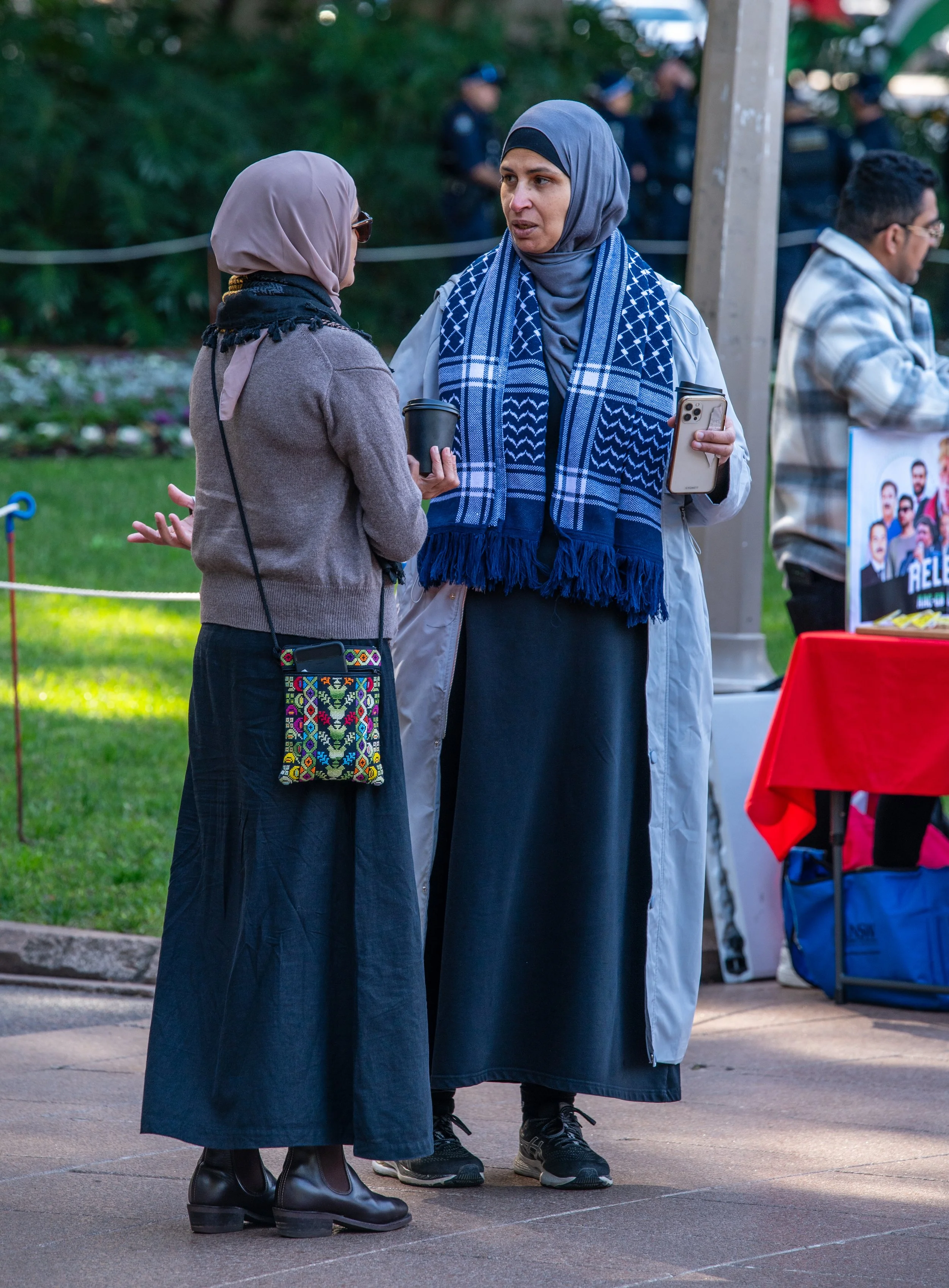 Two women having a conversation outdoors, both wearing hijabs and modest clothing, with one holding a coffee cup and the other holding a phone, while people and a table with a red tablecloth are visible in the background.