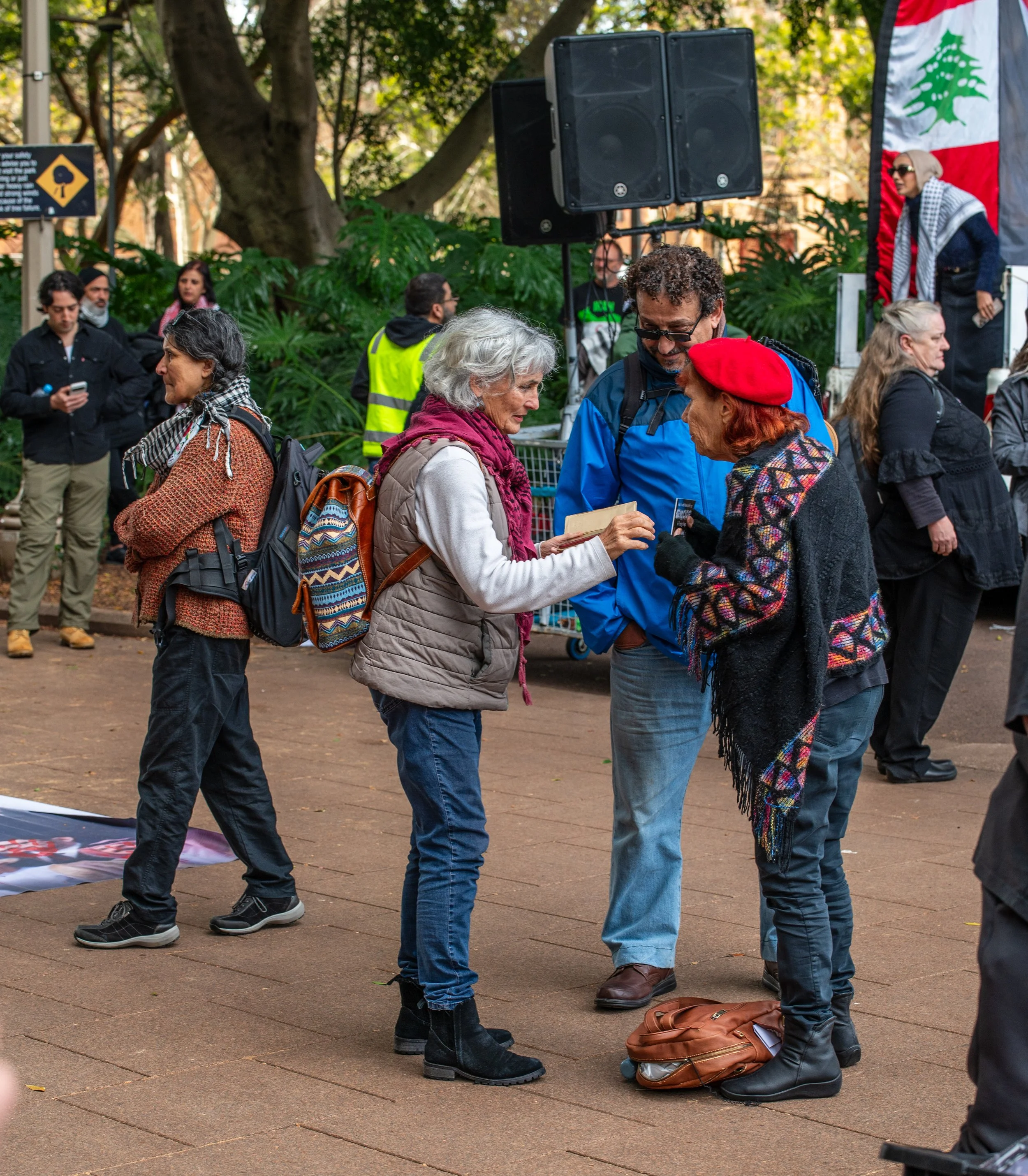 People gathered at an outdoor event, engaging in conversations, with a woman in a red beret showing something on her phone to a man, in front of a Lebanese flag and large speakers.