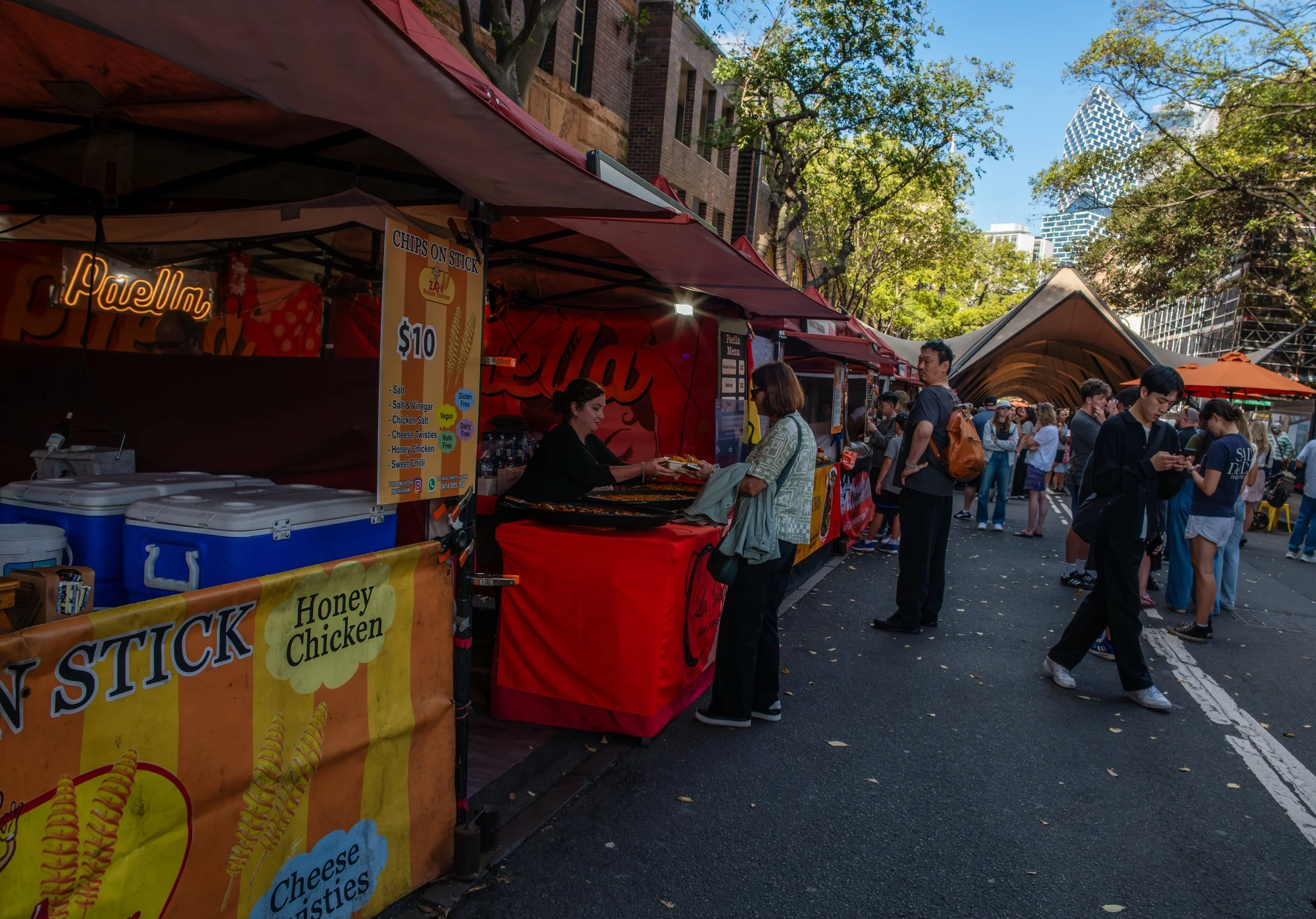 Street food market with a food stall selling honey chicken on stick, surrounded by people walking and ordering, with trees and tall buildings in the background on a sunny day.