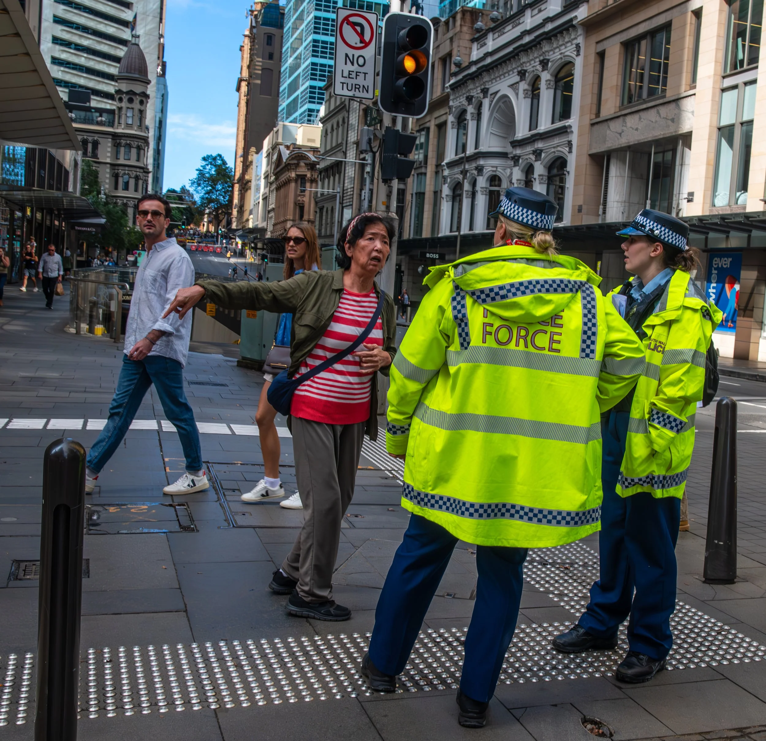 Two police officers in bright yellow jackets talking to a woman on a city sidewalk. The woman is gesturing with her arm as pedestrians pass by. The background features tall buildings and a traffic light showing yellow.