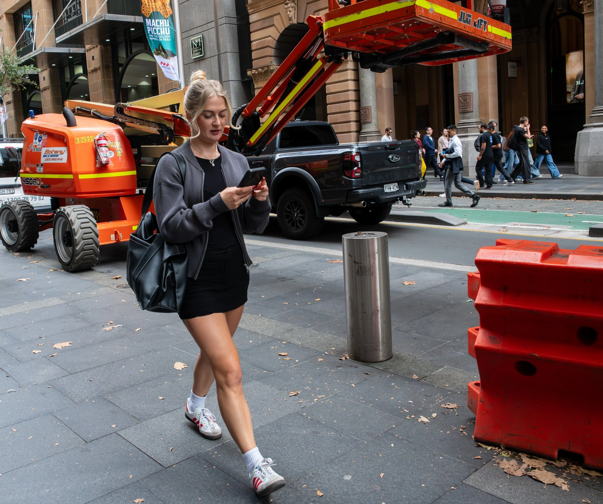 A young woman walking on city sidewalk, looking at her phone, with construction equipment and pedestrians in the background.