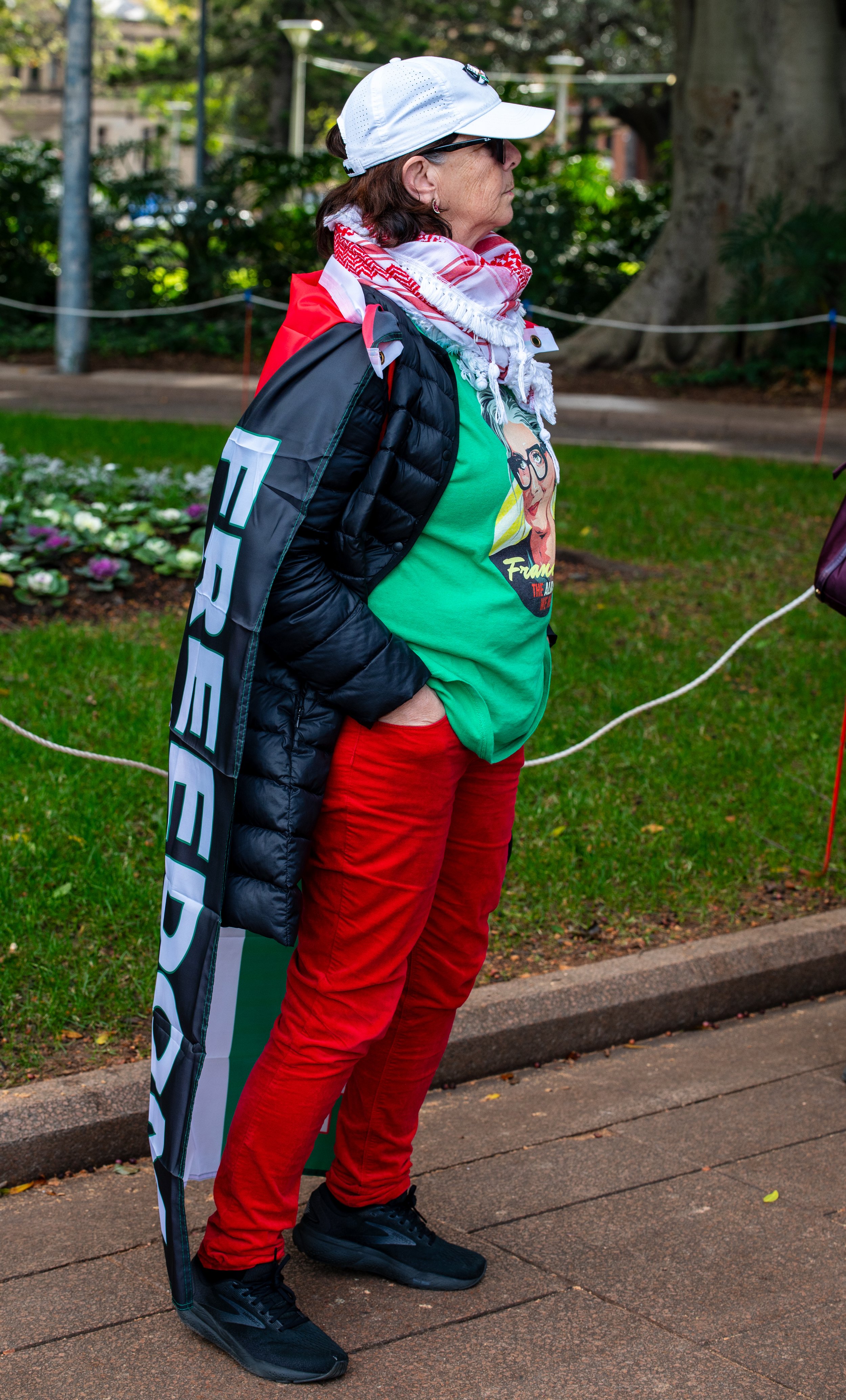 A woman standing outdoors near a garden with flowers and trees, dressed in a white cap, sunglasses, a black puffer jacket, red pants, and a green T-shirt with a cartoon graphic, carrying a flag and wearing a red and white scarf.