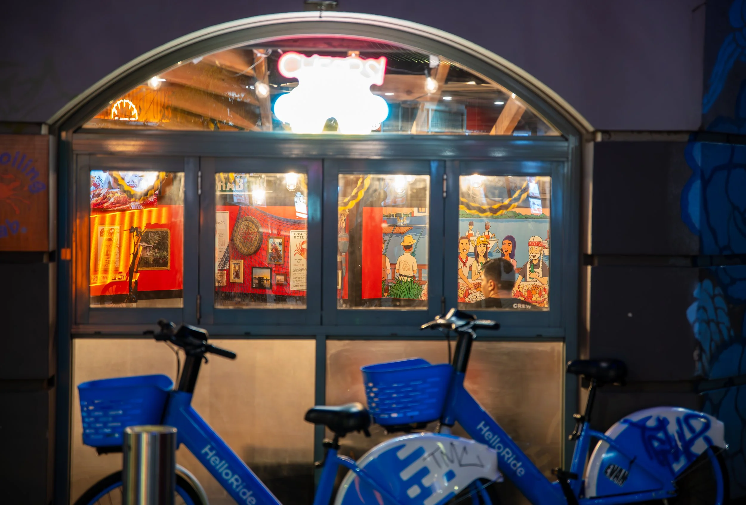 Nighttime view of a small food stand with colorful interior decorations and illustrations, two blue HelloRide bikes parked in front.