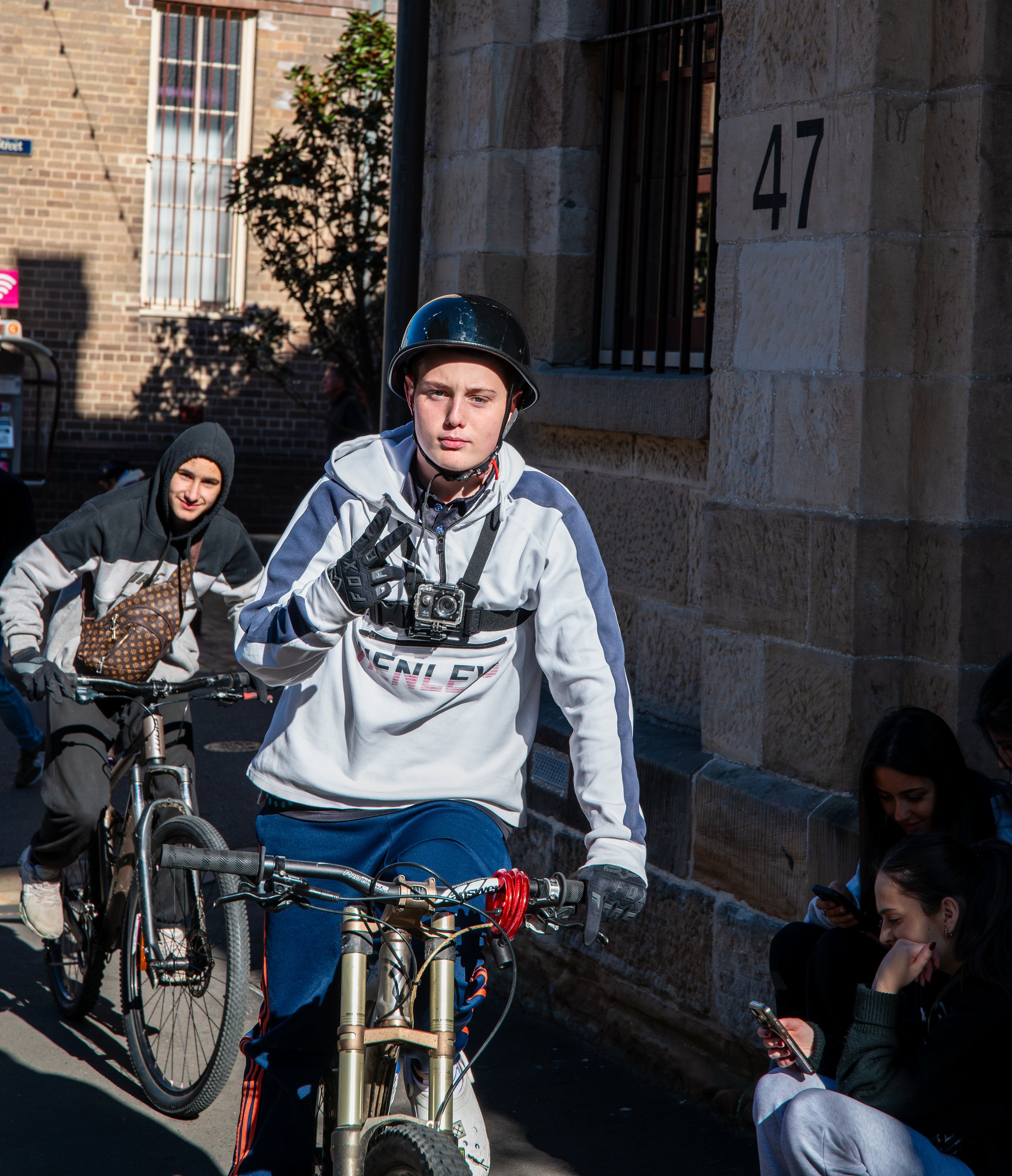 Young man wearing a helmet and gloves riding a mountain bike, making a peace sign with his fingers, with two other cyclists and two women sitting on the sidewalk nearby, in an urban street setting.