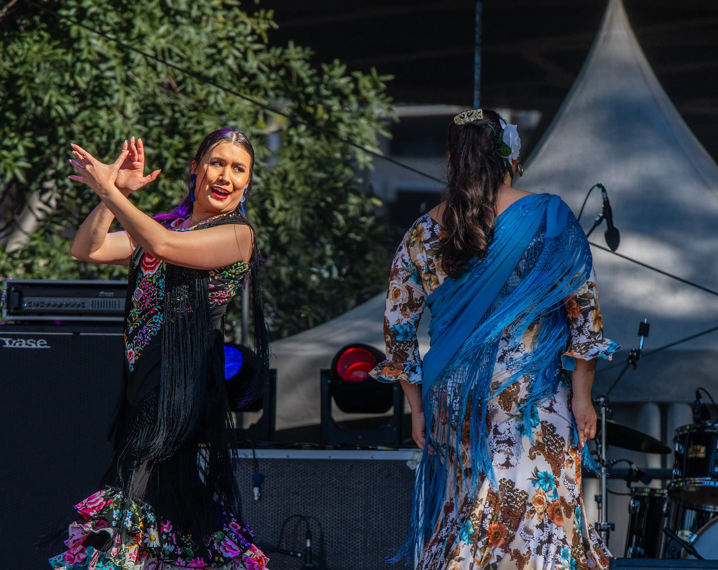 Two women in colorful traditional Mexican dresses perform a dance outdoors on stage with musical equipment in the background.