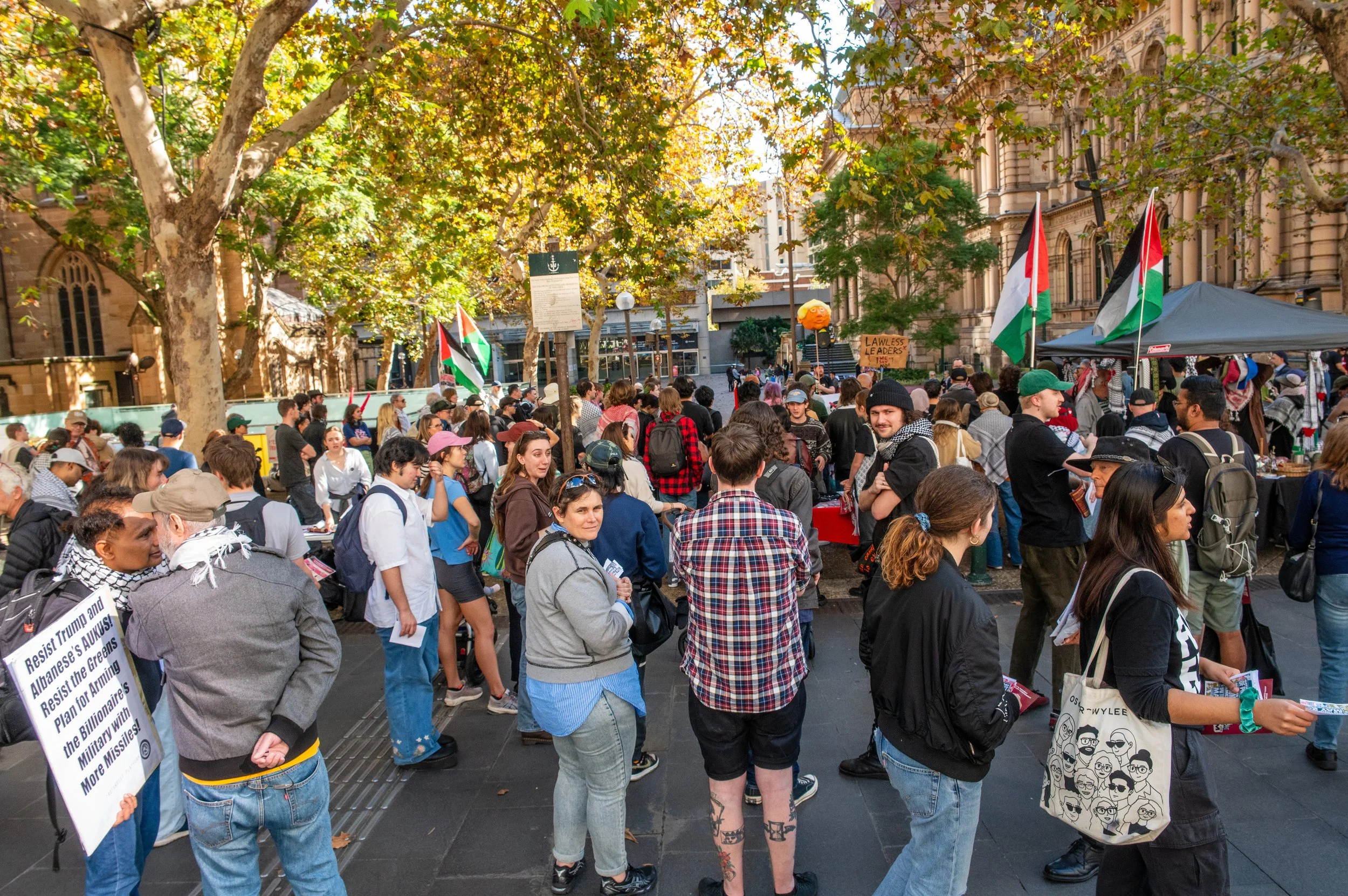Crowd of people gathered outdoors in protest or demonstration, with some holding flags and signs, in a city square shaded by large trees with autumn leaves.