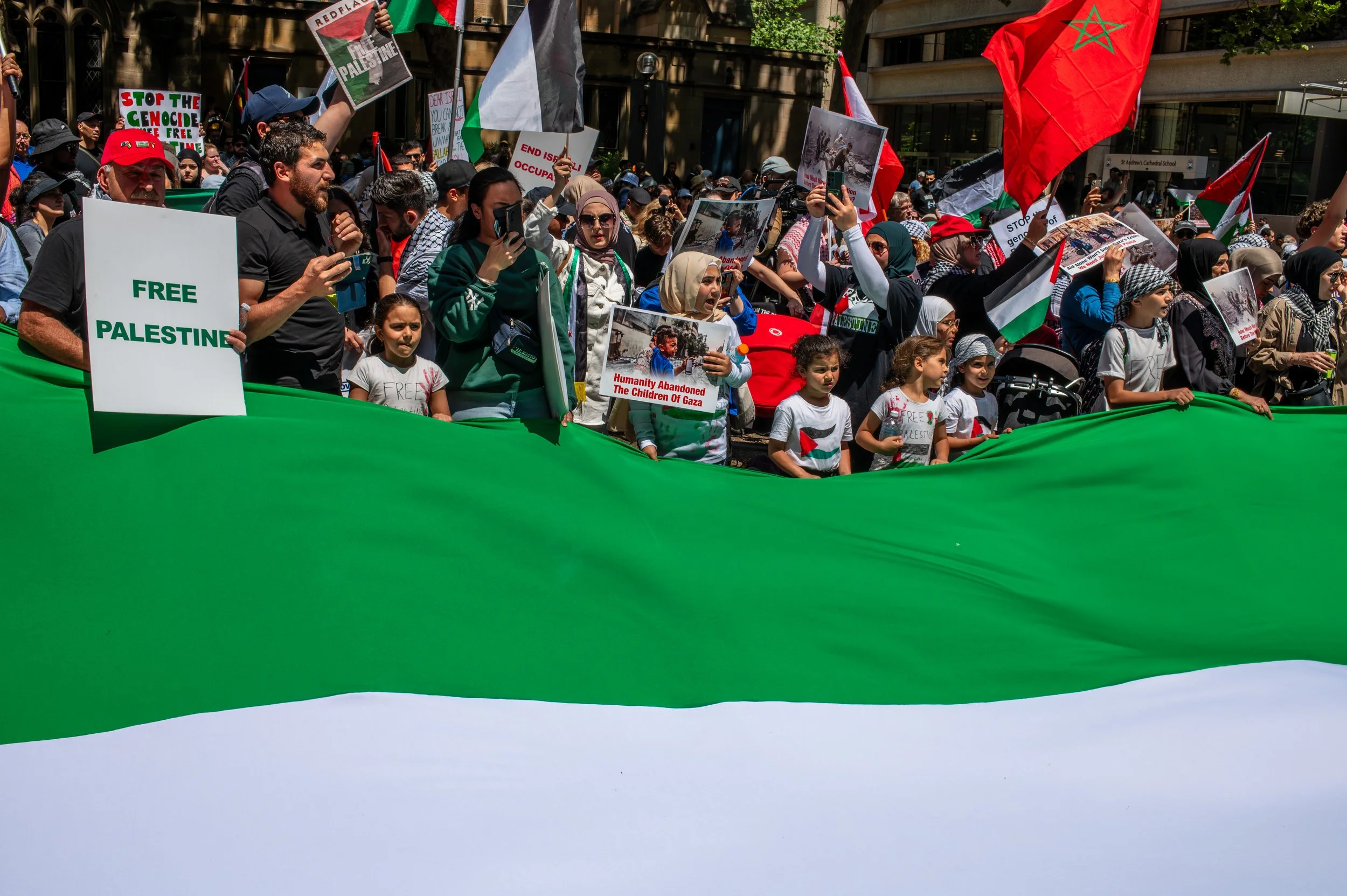 A large group of people protesting, many holding signs and Palestinian flags, with some children present. One sign reads 'Free Palestine.' Others display images and messages in support of Palestine, and a large Palestinian flag is visible among the crowd.