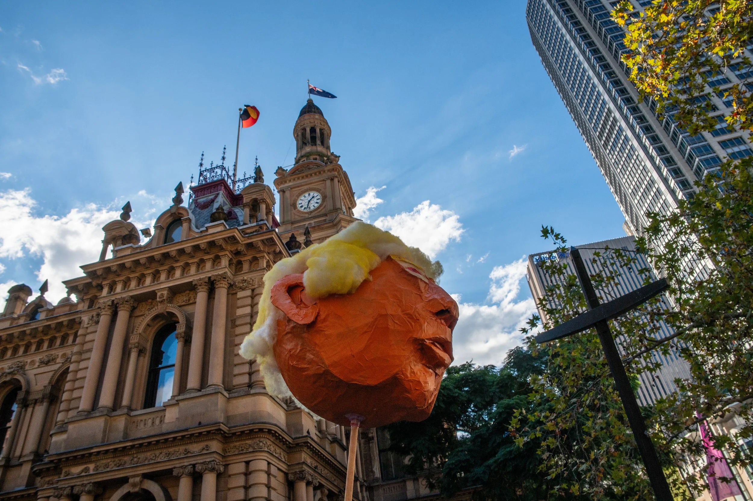A large paper mâché sculpture of a human head with light-colored hair and orange face, held on a stick, set against historic and modern buildings under a partly cloudy sky during daytime.