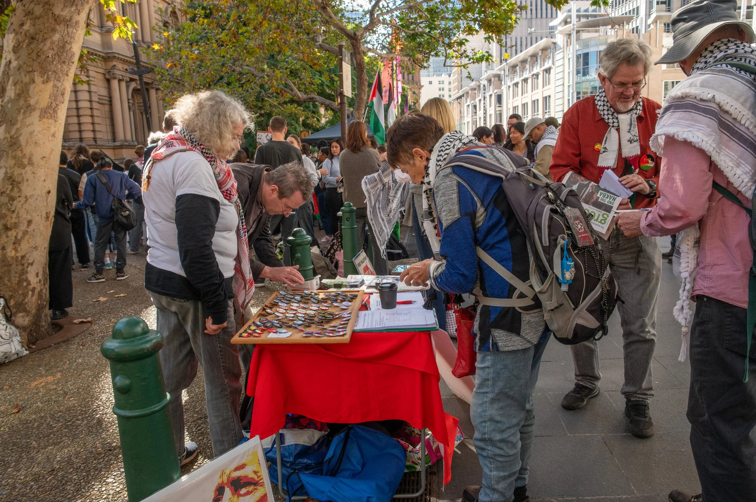 People browse an outdoor market stall with badges or pins on a table, with others looking at pamphlets and flyers, and flags in the background on a city street.