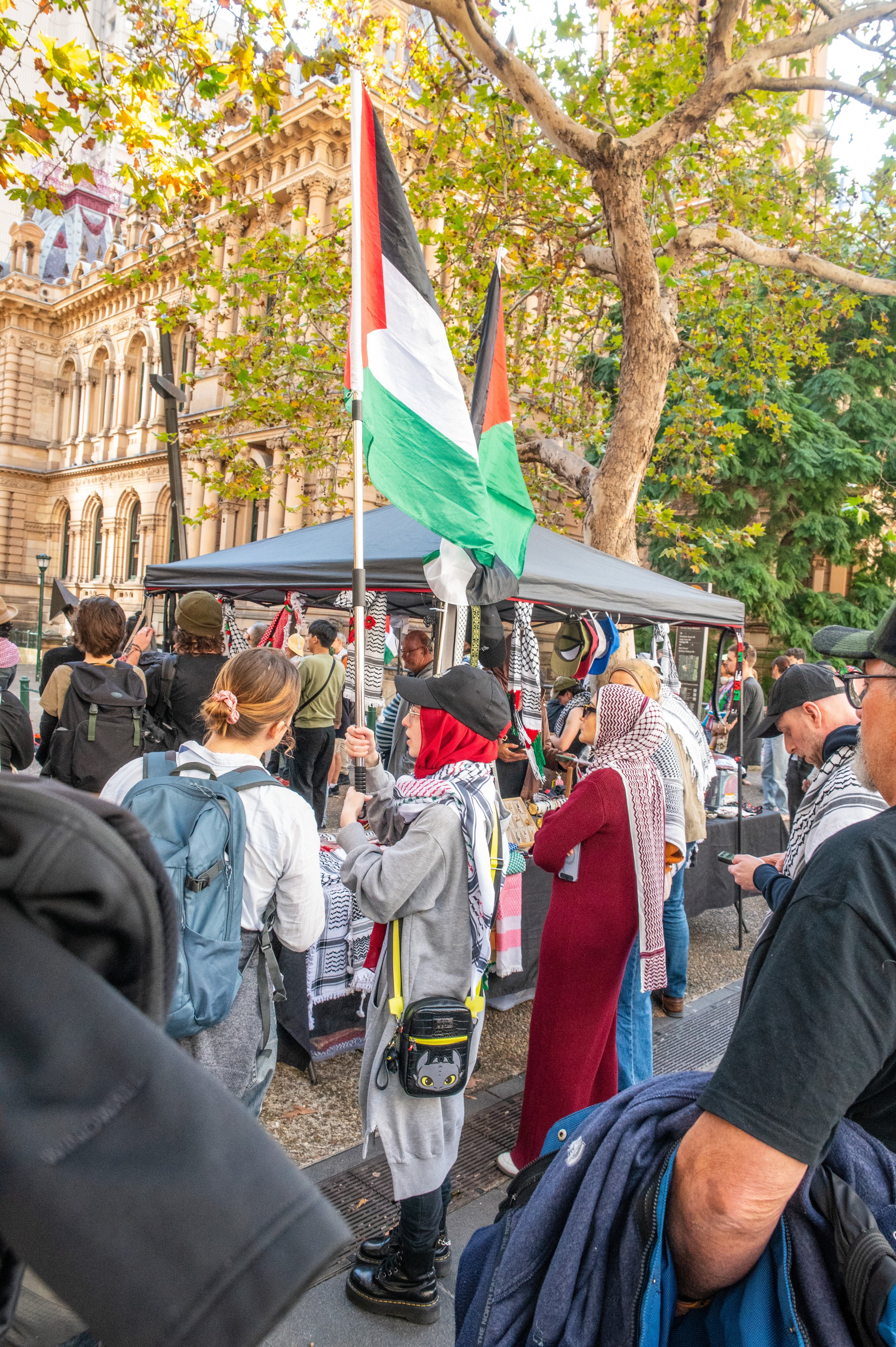 People gathered at a street market with Palestinian flags, some wearing traditional headscarves, and a historic building in the background.