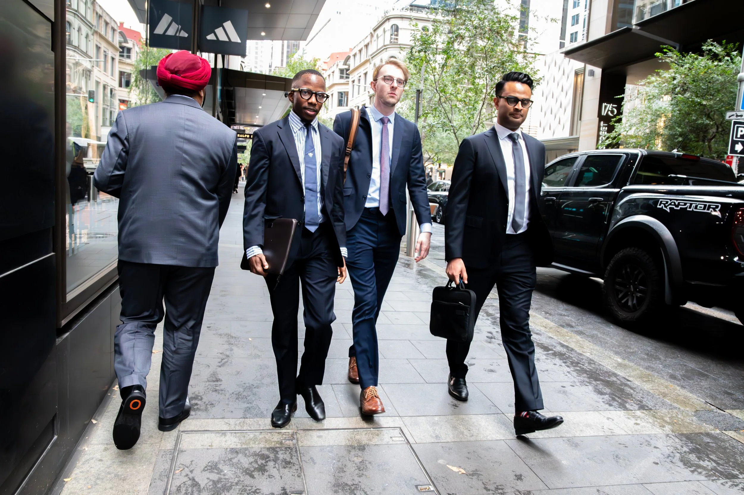 Four men in business suits walking on a city sidewalk, two carrying briefcases and one with a laptop, with office buildings and parked trucks in the background.