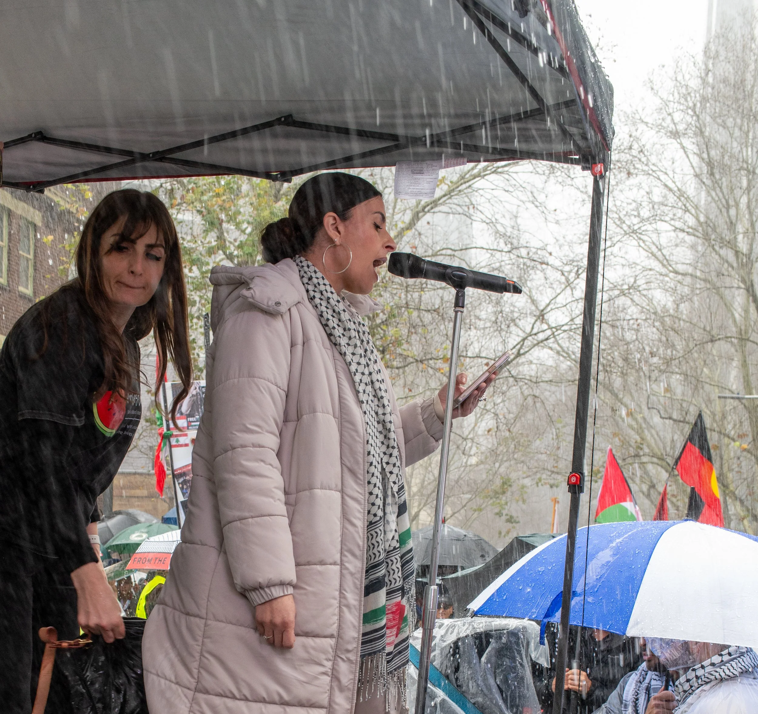 Woman speaking at an outdoor event, holding a phone, under a canopy in the rain. Another woman stands nearby. People with umbrellas are visible in the background, some carrying flags.