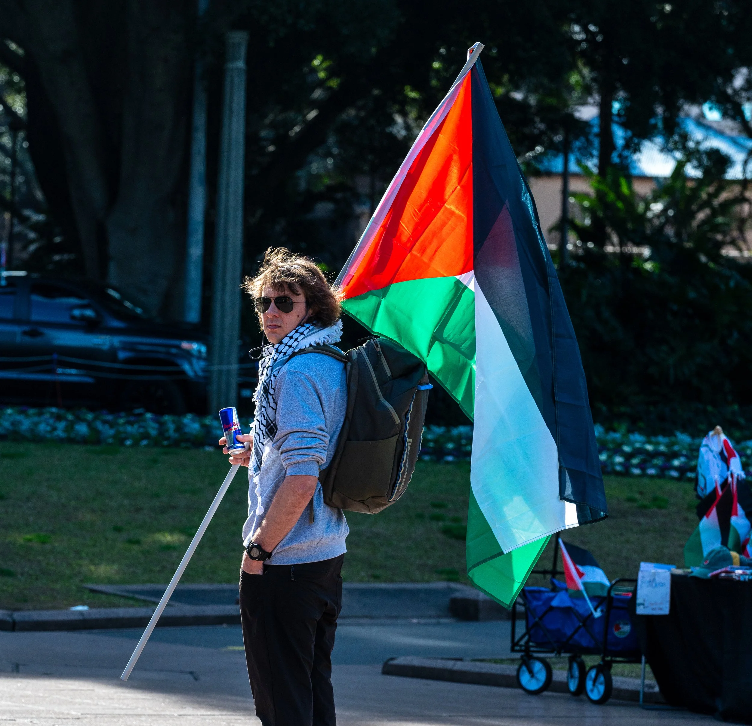 A person holding a Palestinian flag and a can of Red Bull, standing outdoors on a street with trees and parked cars in the background.