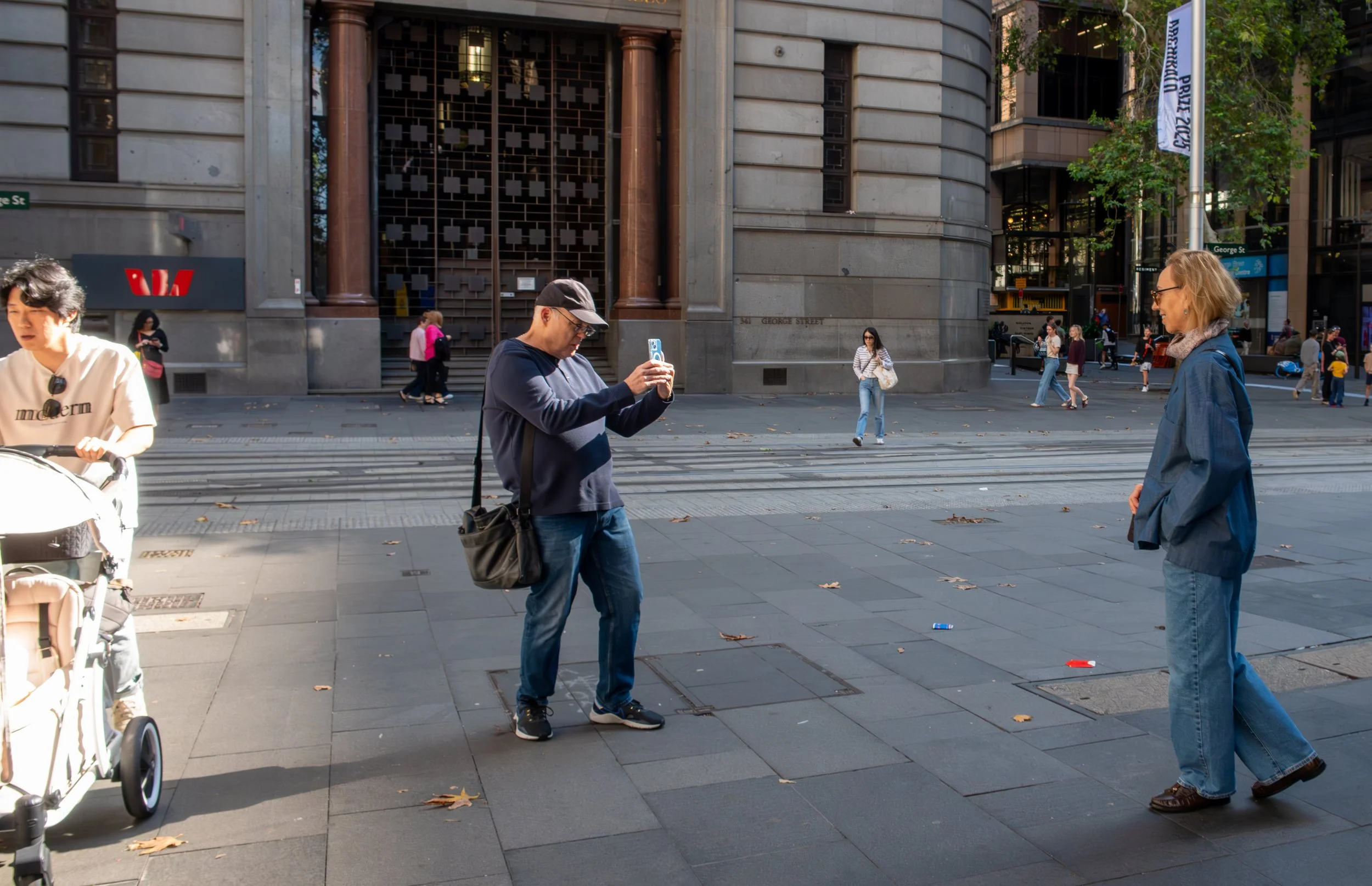 Two people standing on a city sidewalk, one taking a photo of the other. Several pedestrians walk in the background near a large stone building with a water emblem sign.