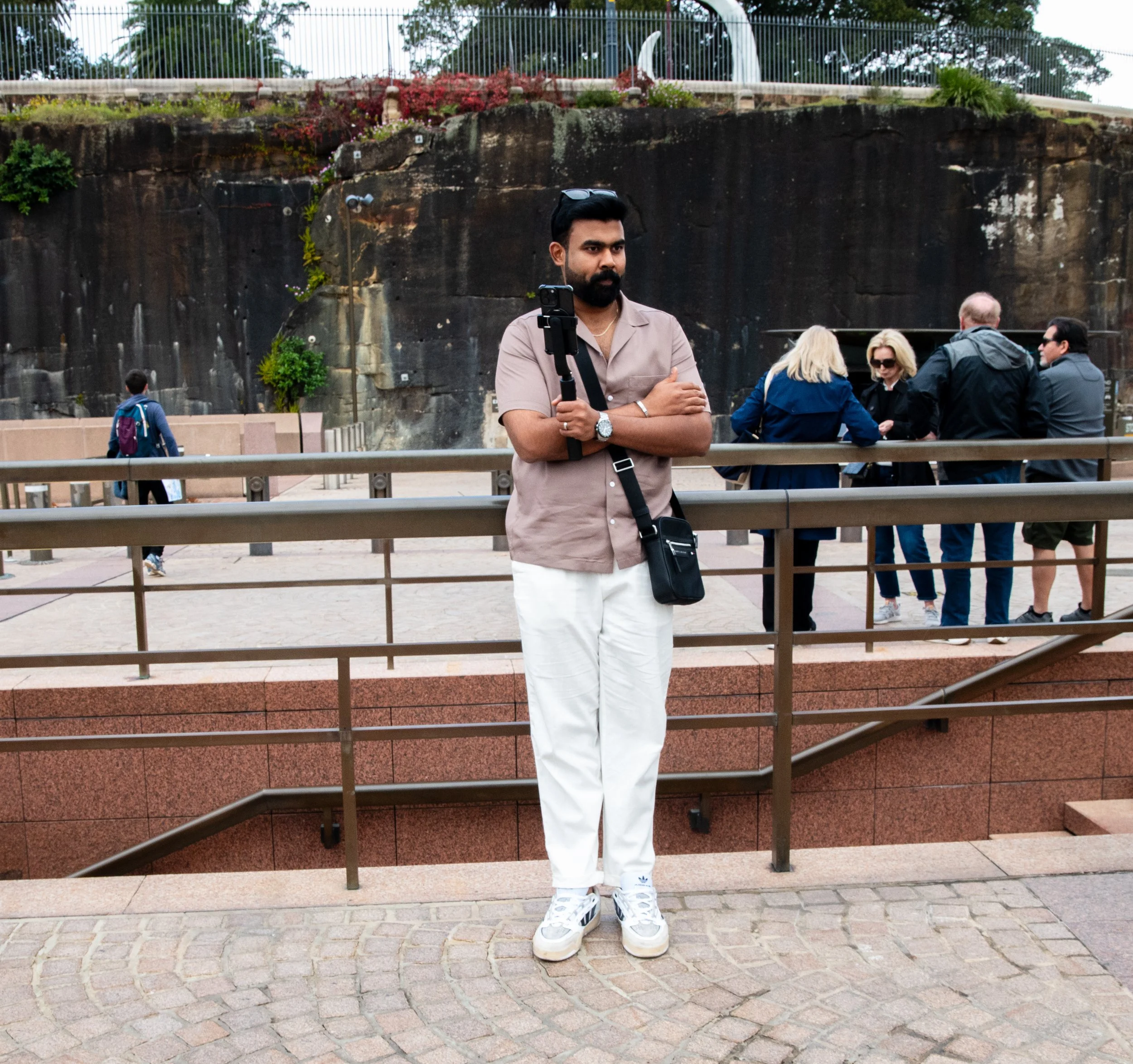 A man standing in front of a metal railing at an outdoor venue with a group of people gathered behind him. He is holding a tripod with a camera or phone mounted on it, wearing a beige shirt, white pants, and sneakers, with sunglasses on his head. There is a rocky wall with plants and trees in the background.