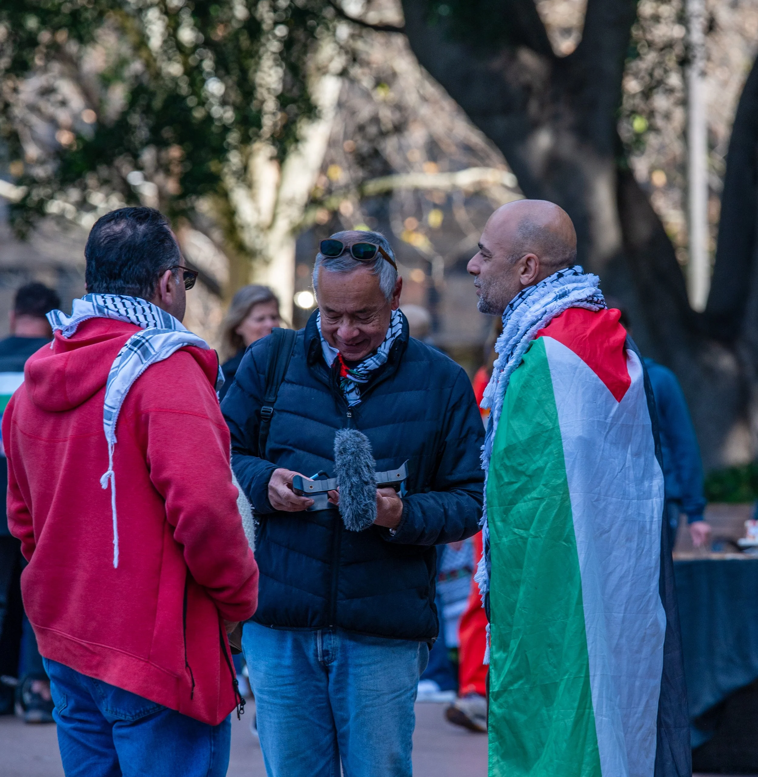 Three men are standing together outdoors, engaged in conversation. The man on the right is wrapped in a flag with red, green, white, and black colors. The man in the middle is holding a drone and appears to be showing it to the others, while the man on the left is wearing a red hoodie. Trees with bare branches are in the background.