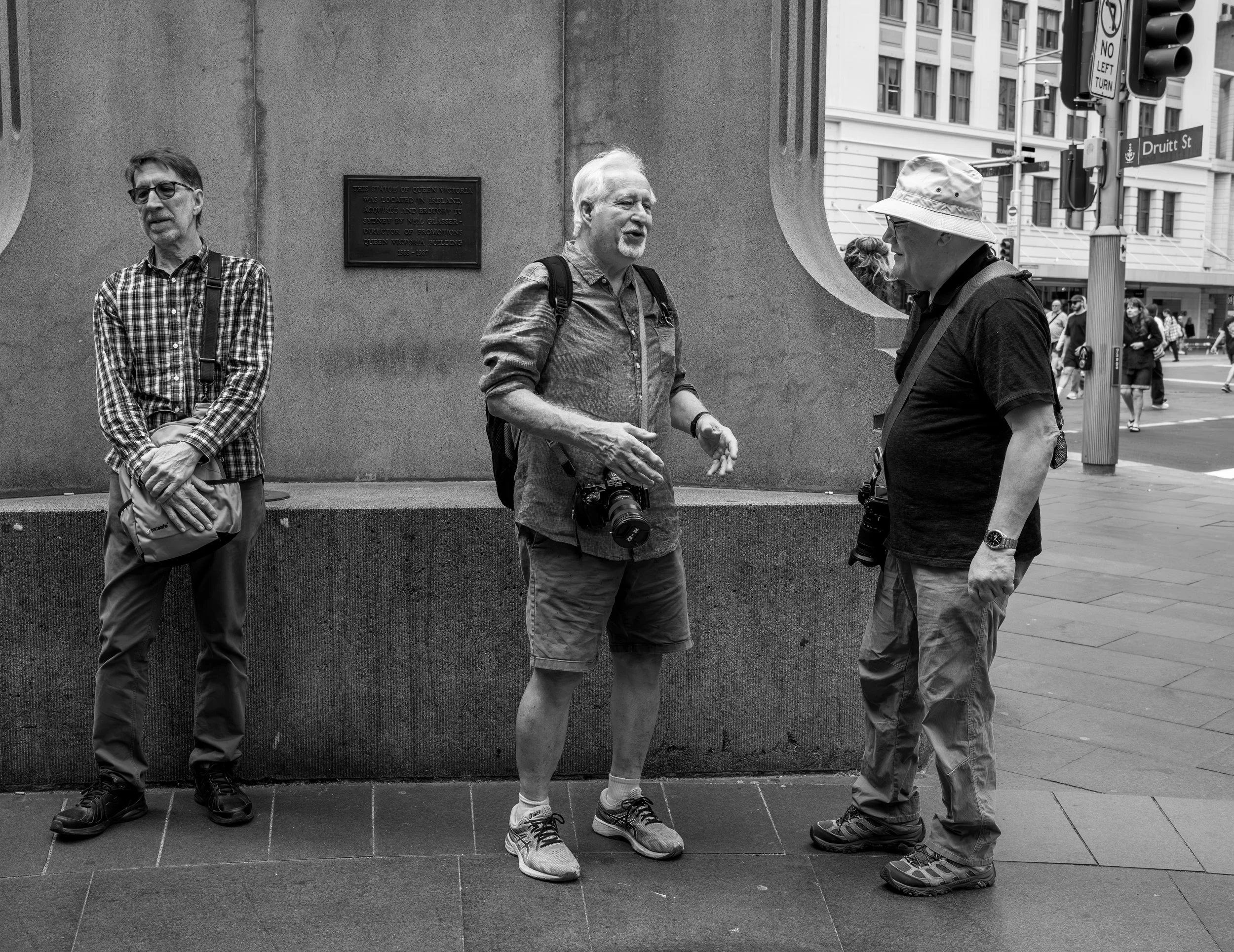 Three men with cameras and backpacks standing on a city sidewalk, having a conversation in front of a stone wall with a plaque.