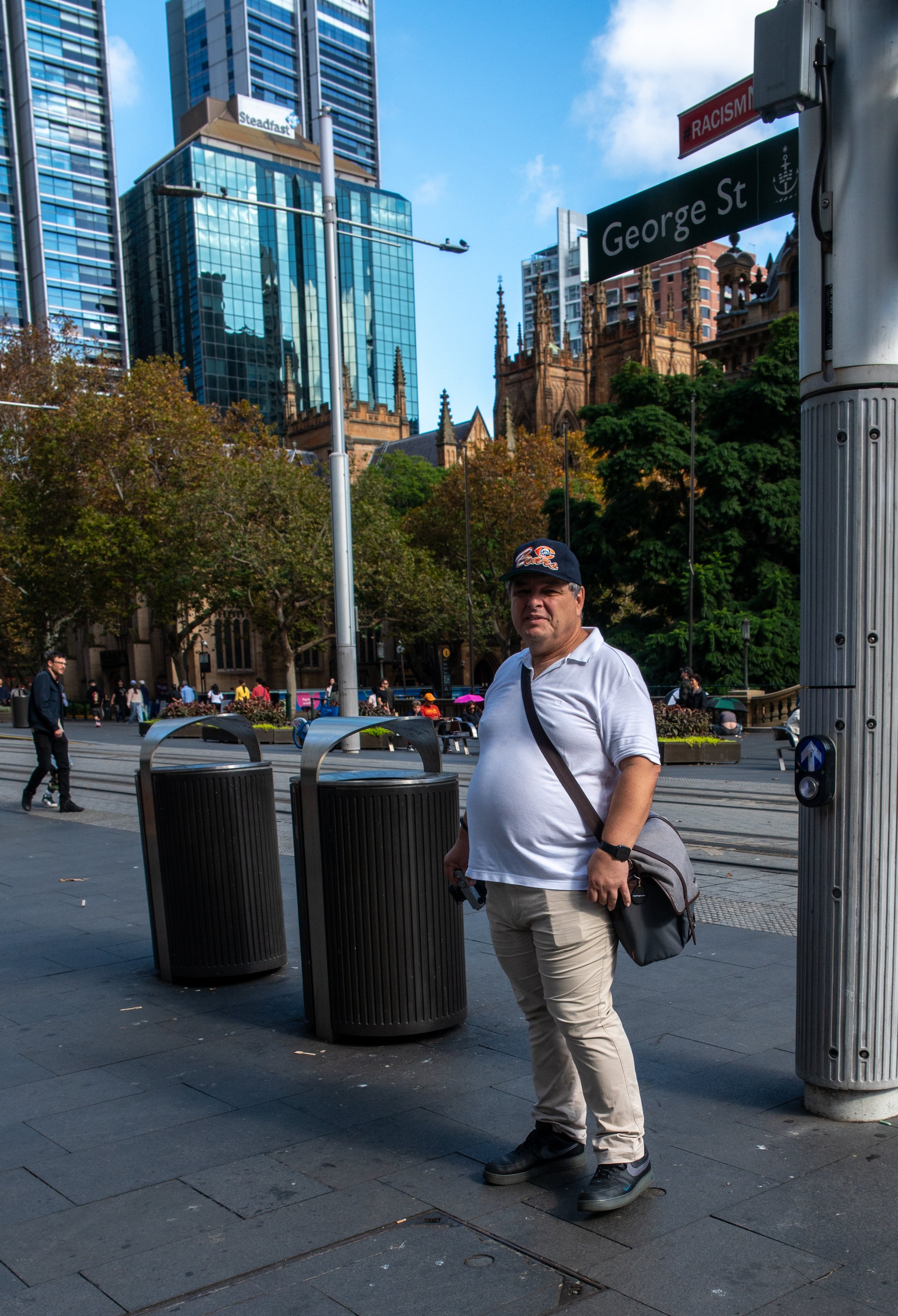 A man in a white polo shirt, beige pants, and a baseball cap stands on a city street near trash cans and street signs with high-rise buildings and a church in the background.