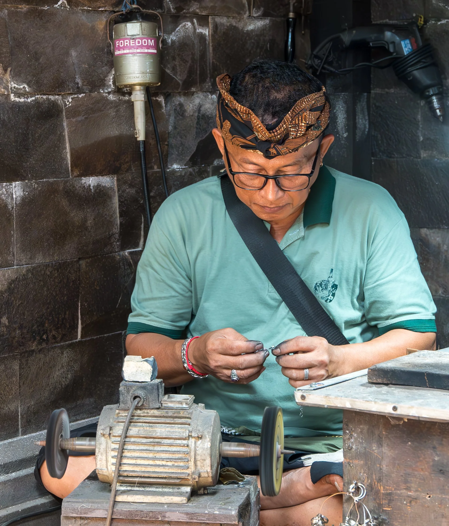 A man working with jewelry or small metal objects, wearing glasses, a headscarf, and a green shirt, sitting at a workbench with tools and equipment around him.