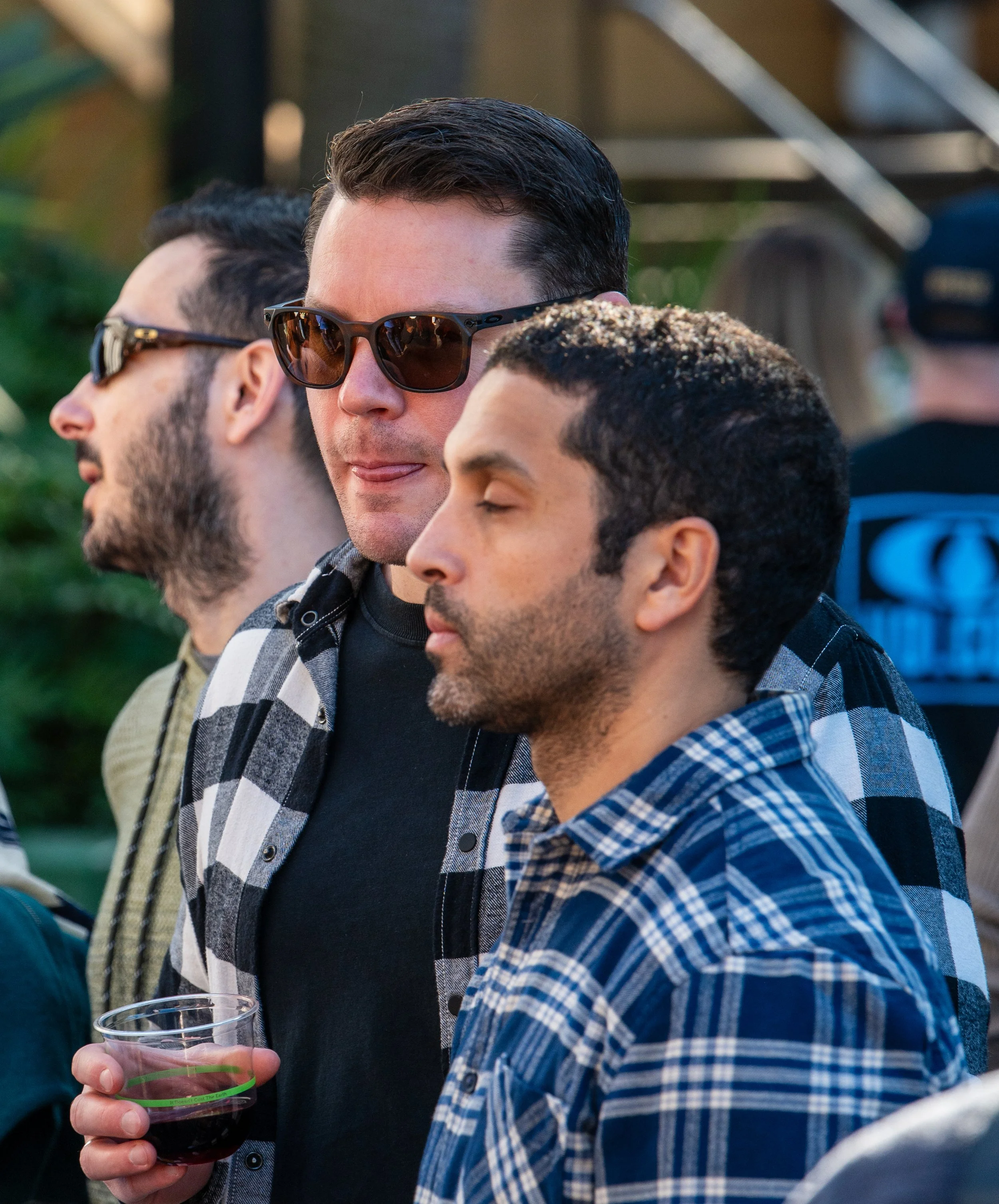 Three men standing in a row outdoors, one wearing sunglasses and holding a glass of wine, all appear to be at a social event or gathering.