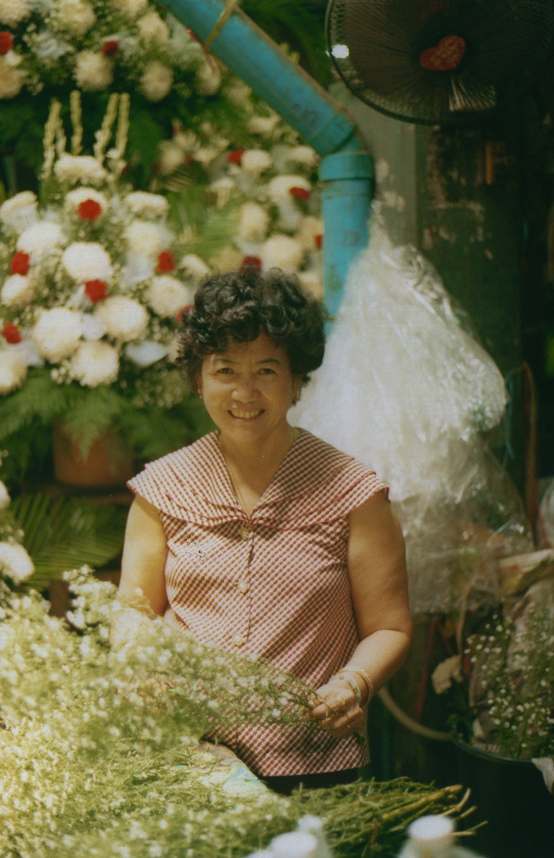 A smiling woman with short curly hair wearing a red and white checkered sleeveless dress, standing among flower arrangements in a flower shop or market.