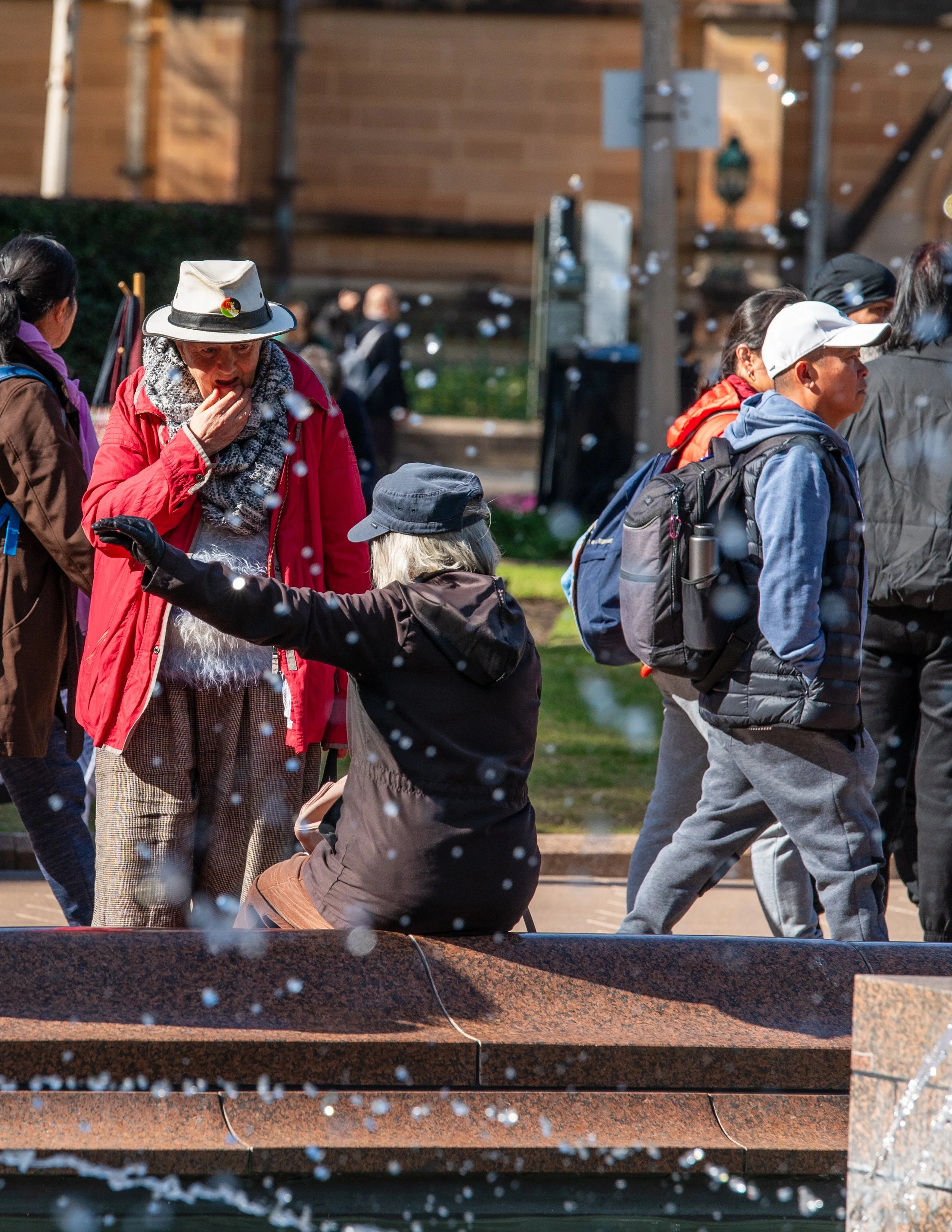 A group of people gathered outdoors near a fountain, some wearing backpacks, with a woman in a red coat and white hat touching her face, and another woman sitting near the fountain reaching out with her hand.