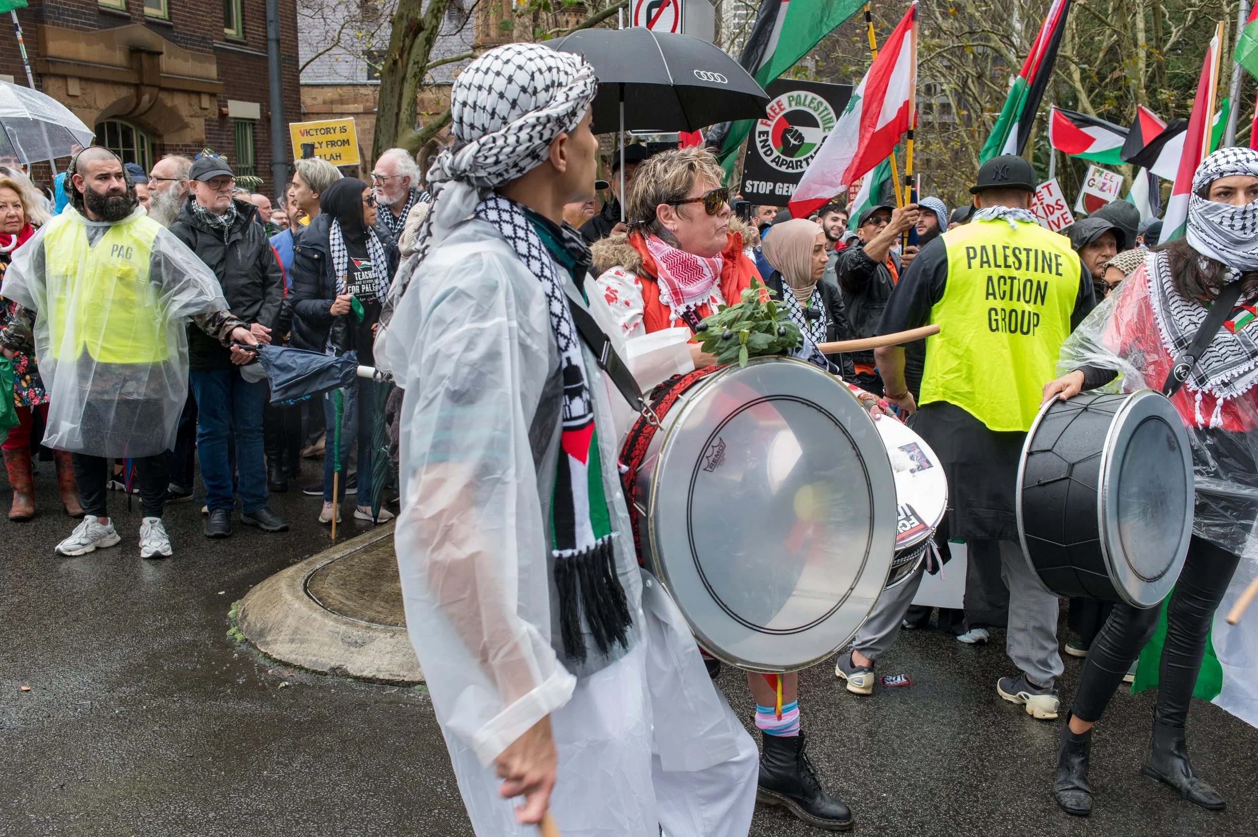 Crowd of people participating in a protest, some playing drums. Many hold Palestinian flags and signs advocating for Palestine, with rain-soaked streets and people dressed warmly.
