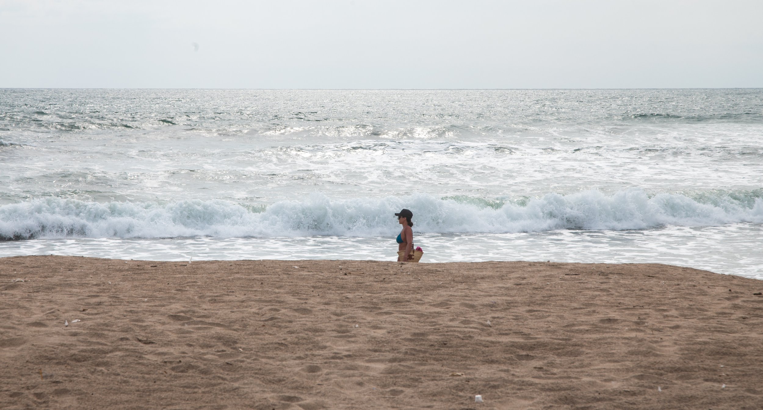 A woman wearing a blue top and beige shorts walking along the sandy beach near the water, carrying a basket.