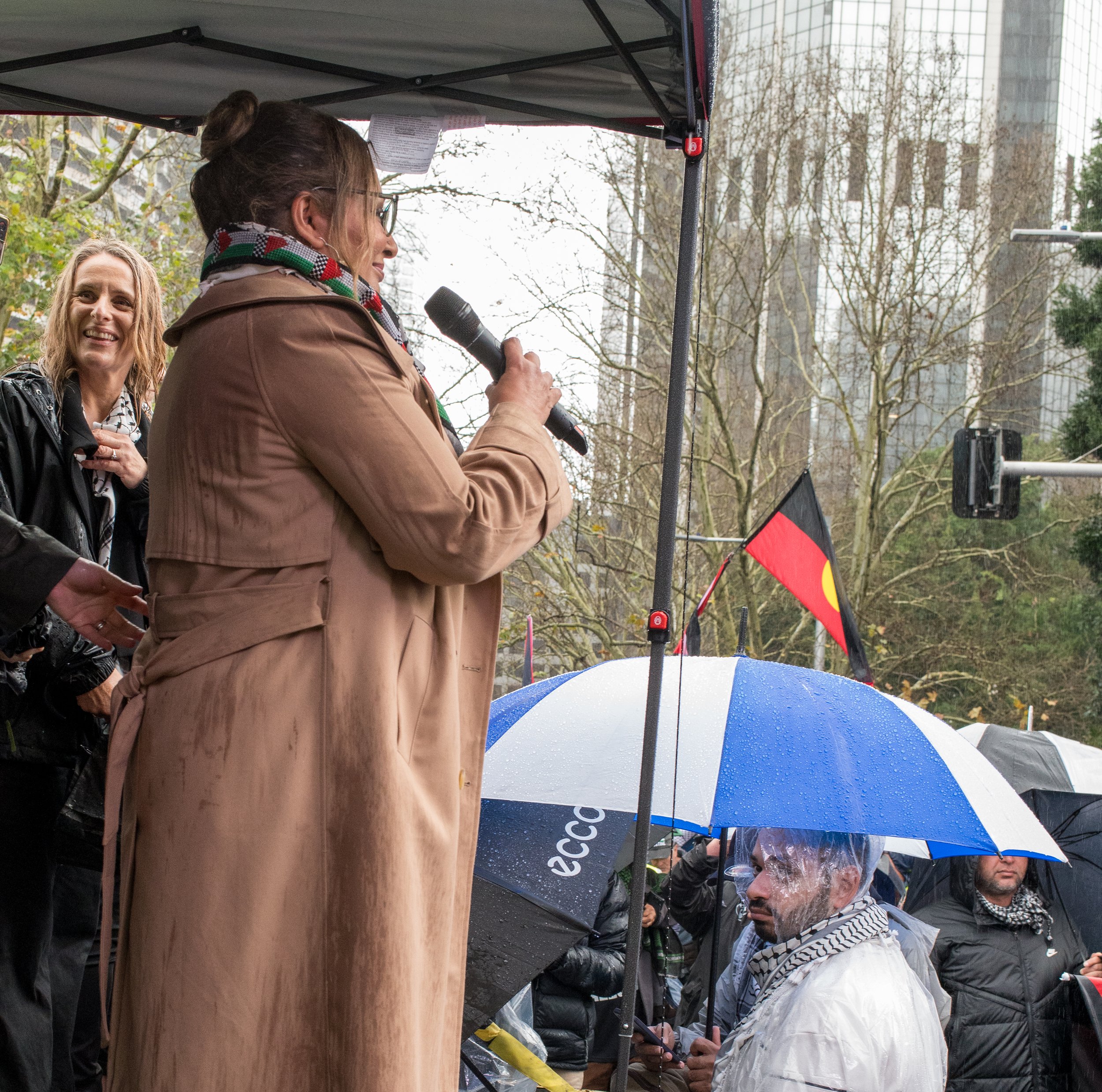 A woman speaking into a microphone at a protest or rally outdoors in rainy weather, with people holding umbrellas and a Native American flag visible in the background.