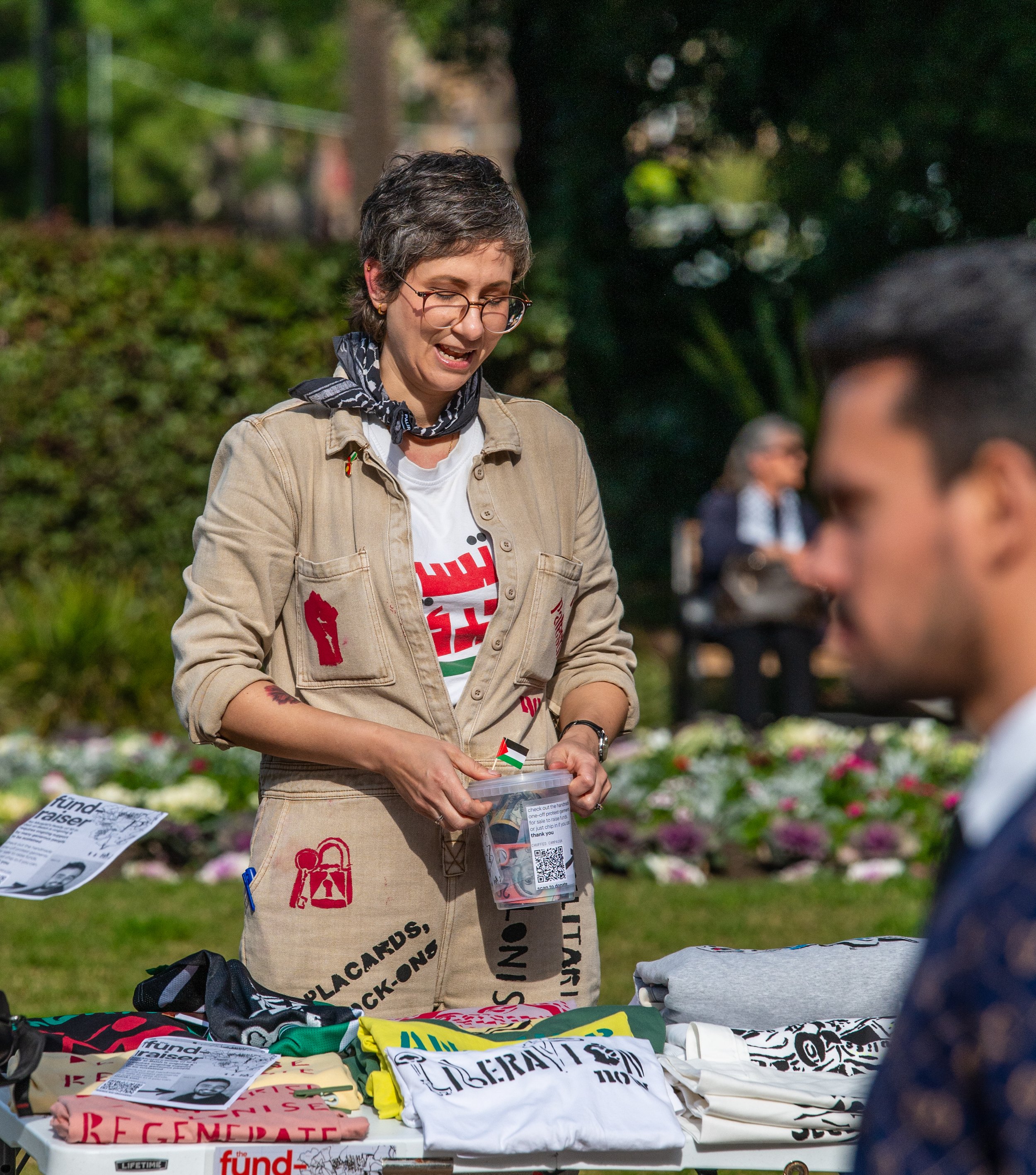 A woman with short gray hair and glasses is standing outdoors behind a table of folded T-shirts. She is smiling and holding a small item in her hands. The woman is wearing a khaki jacket with red patches, a white T-shirt, and a black bandana around her neck. There are people and trees in the background.