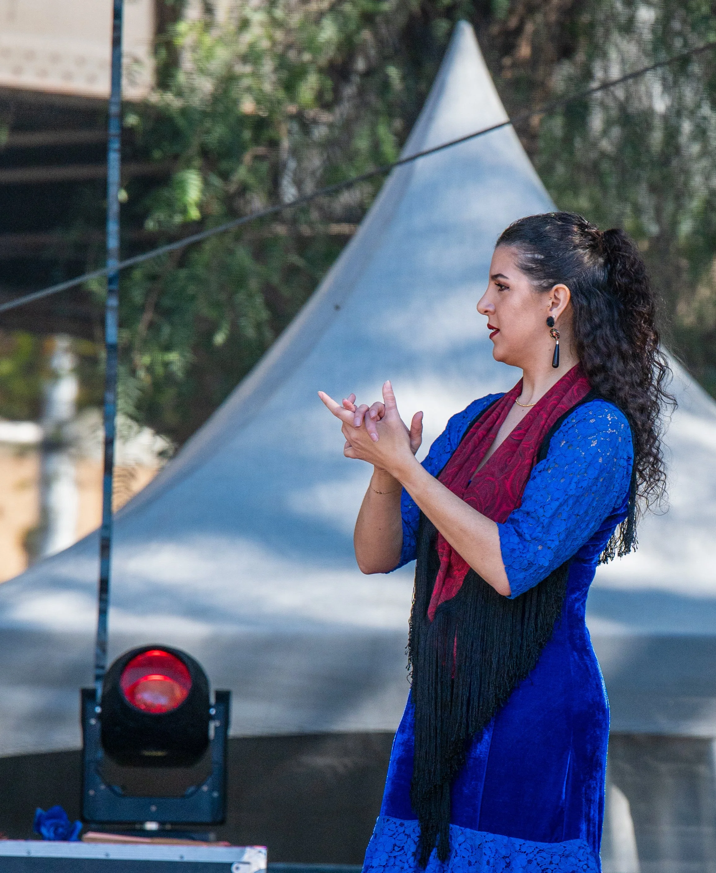 A woman with curly dark hair wearing a blue dress and red scarf, standing outdoors in front of a tent, clasping her hands together.