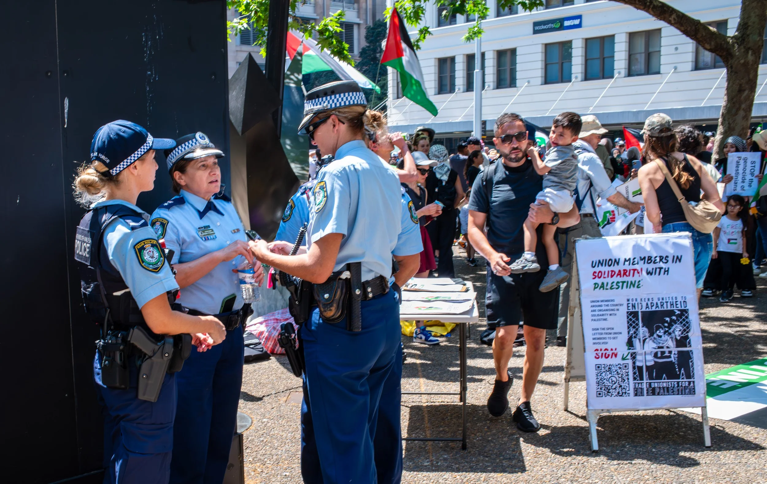 Group of police officers in uniform talking to a man and a woman at a protest, with a sign supporting Palestine and solidarity messages, and people holding flags and signs in the background.