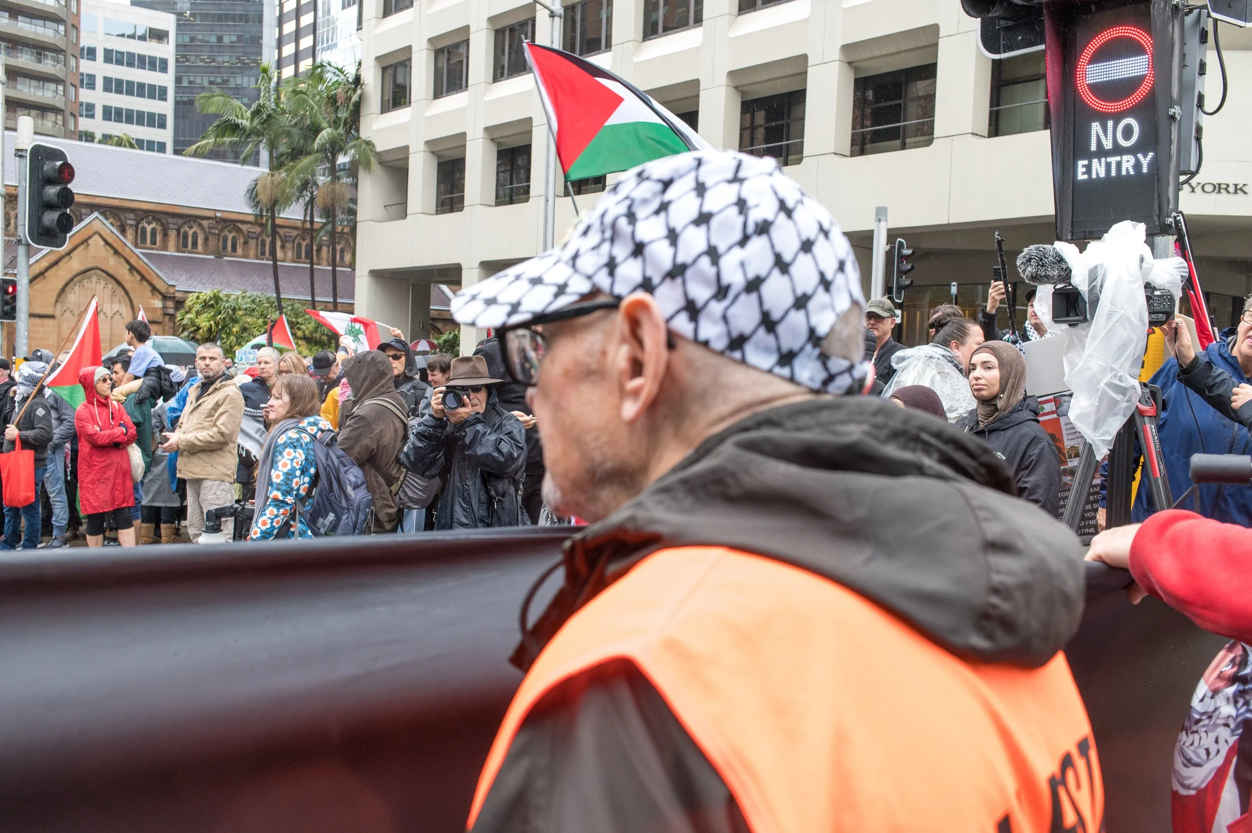 Crowd of people protesting on a city street, some holding Palestinian flags, with a person wearing a black and white checkered keffiyeh pattern hat and an orange vest in the foreground. Cars and high-rise buildings are visible in the background, along with a traffic light showing a red signal and a sign that says 'No Entry'.