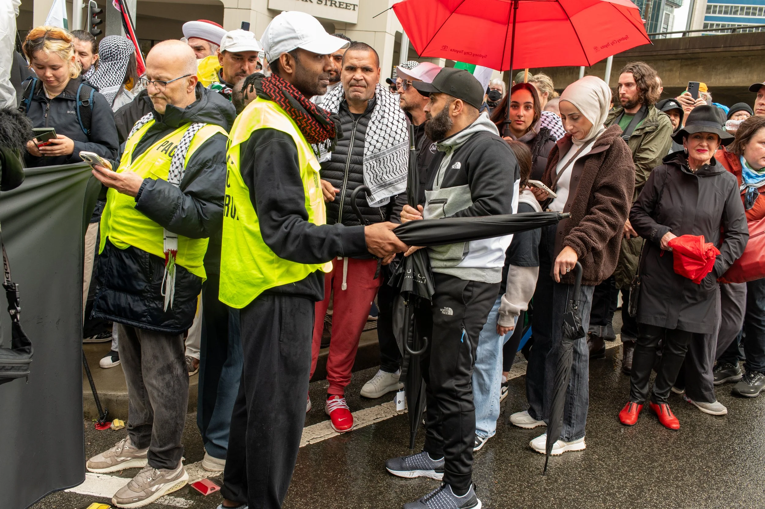 A group of people gathered outdoors on a rainy day, some holding umbrellas, with two individuals in bright yellow vests in the center having a conversation. Others are looking at their phones or observing the scene, and some are wearing scarves or hats.