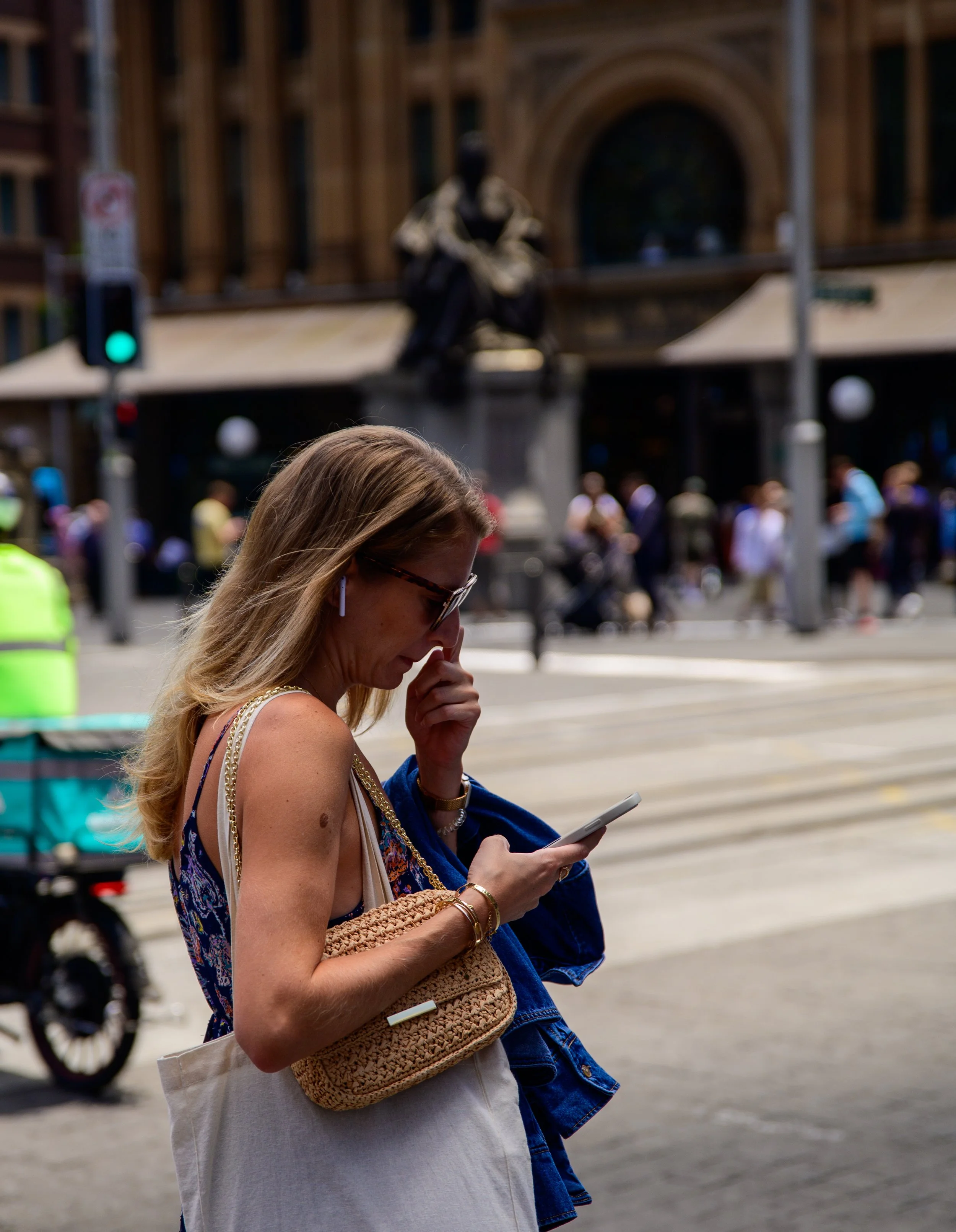 A woman with blonde hair, wearing sunglasses, a tank top, and jewelry, is looking at her phone while holding a woven bag and a blue jacket. She is on a city street with a busy crowd and a large statue of a seated figure in the background.