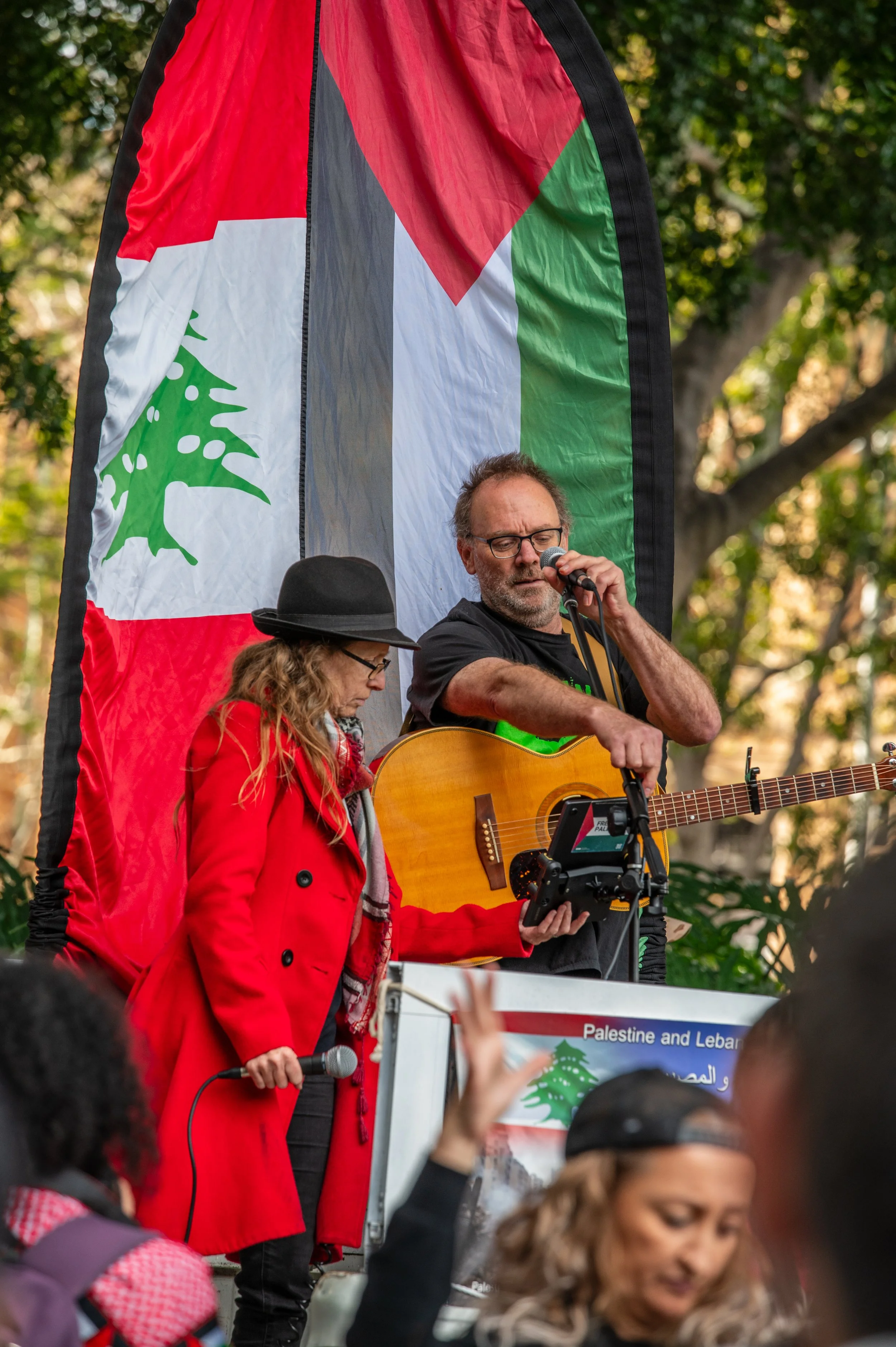 Two performers on stage at a Palestine and Lebanon protest, one woman in a red coat and hat holding a microphone, the other man with glasses playing an acoustic guitar and singing into a microphone. Behind them is a large flag with green, red, black, and white colors, featuring a green cedar tree emblem, set outdoors with trees in the background.