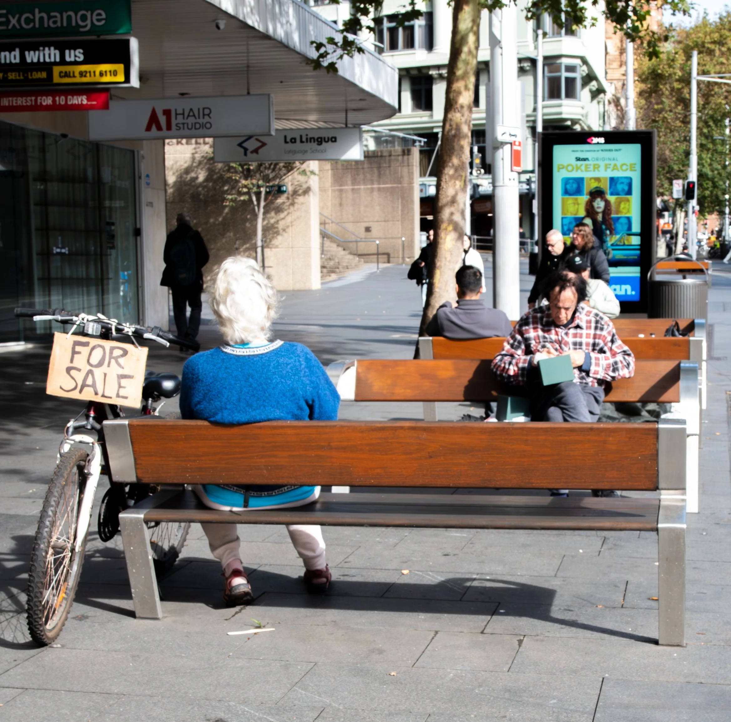 A woman with white hair and a blue sweater sitting on a park bench with a bicycle parked next to her that has a 'For Sale' sign. Behind her, a man with dark curly hair and a checkered shirt is sitting on another bench reading a book. In the background, there are pedestrians, trees, digital advertising boards, and storefronts, suggesting an urban area.