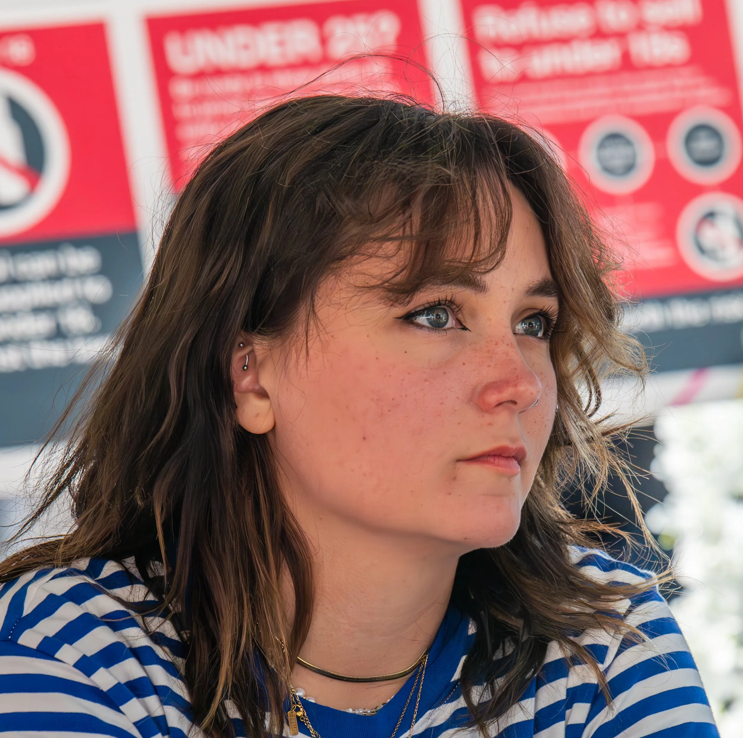 A young woman with wavy brown hair, blue eyes, and freckles, wearing striped blue and white shirt, layered necklaces, and small earrings, looking to her left in front of a red informational sign.