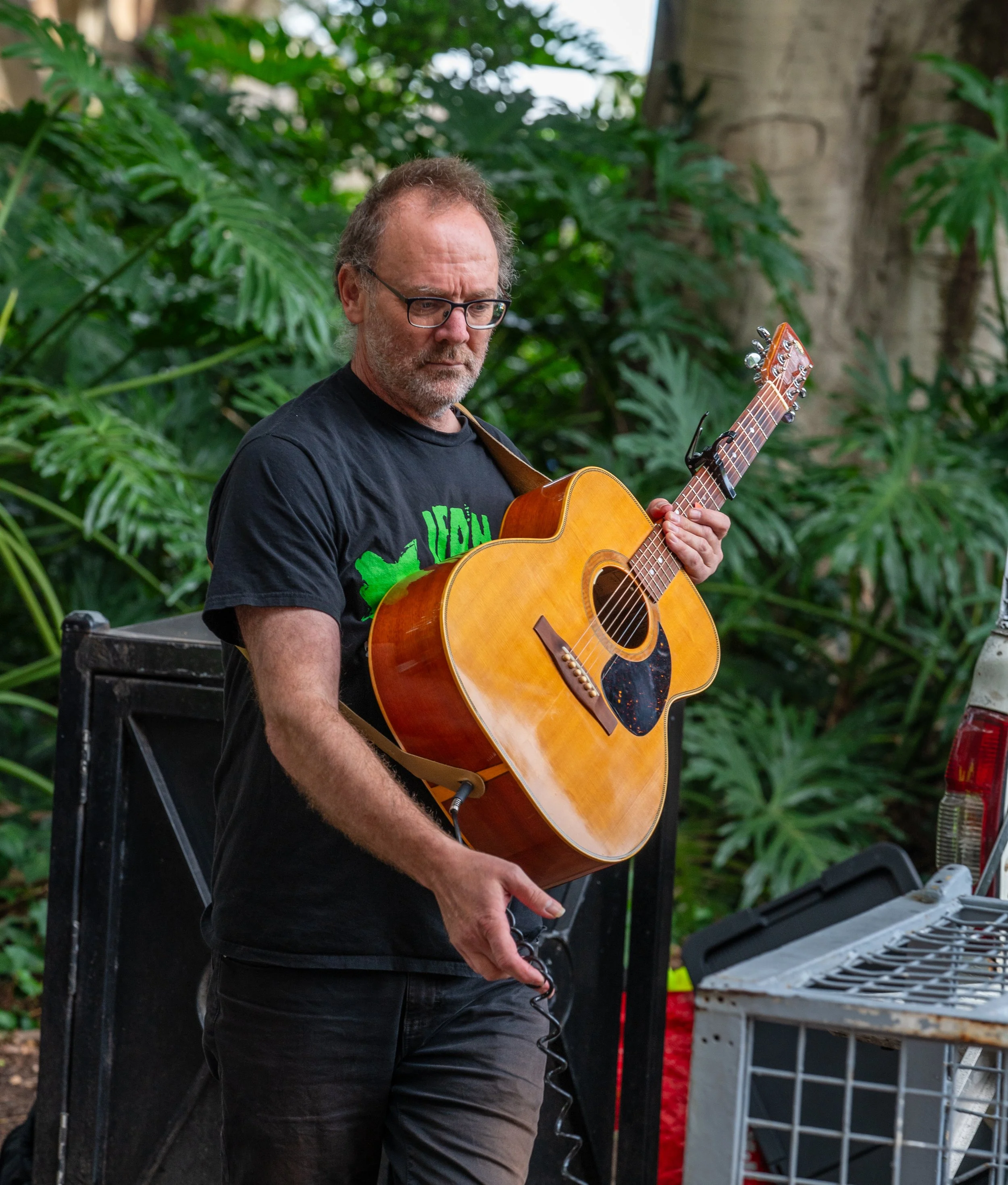 A man with glasses and a beard playing an acoustic guitar outdoors, surrounded by green foliage.