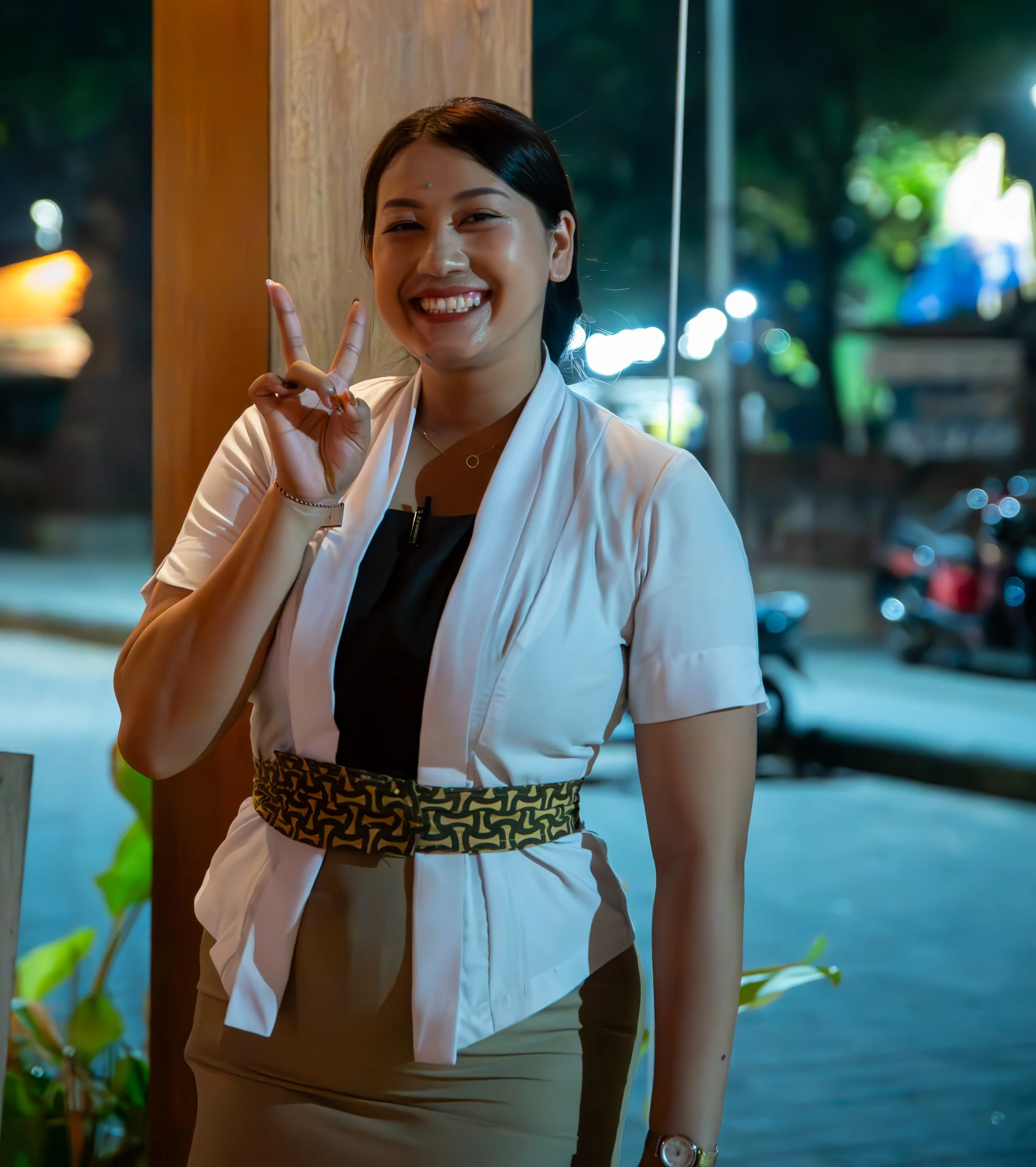 A woman smiling and making a peace sign with her hand, standing near a wooden post outside during evening or night, with blurred lights and greenery in the background.
