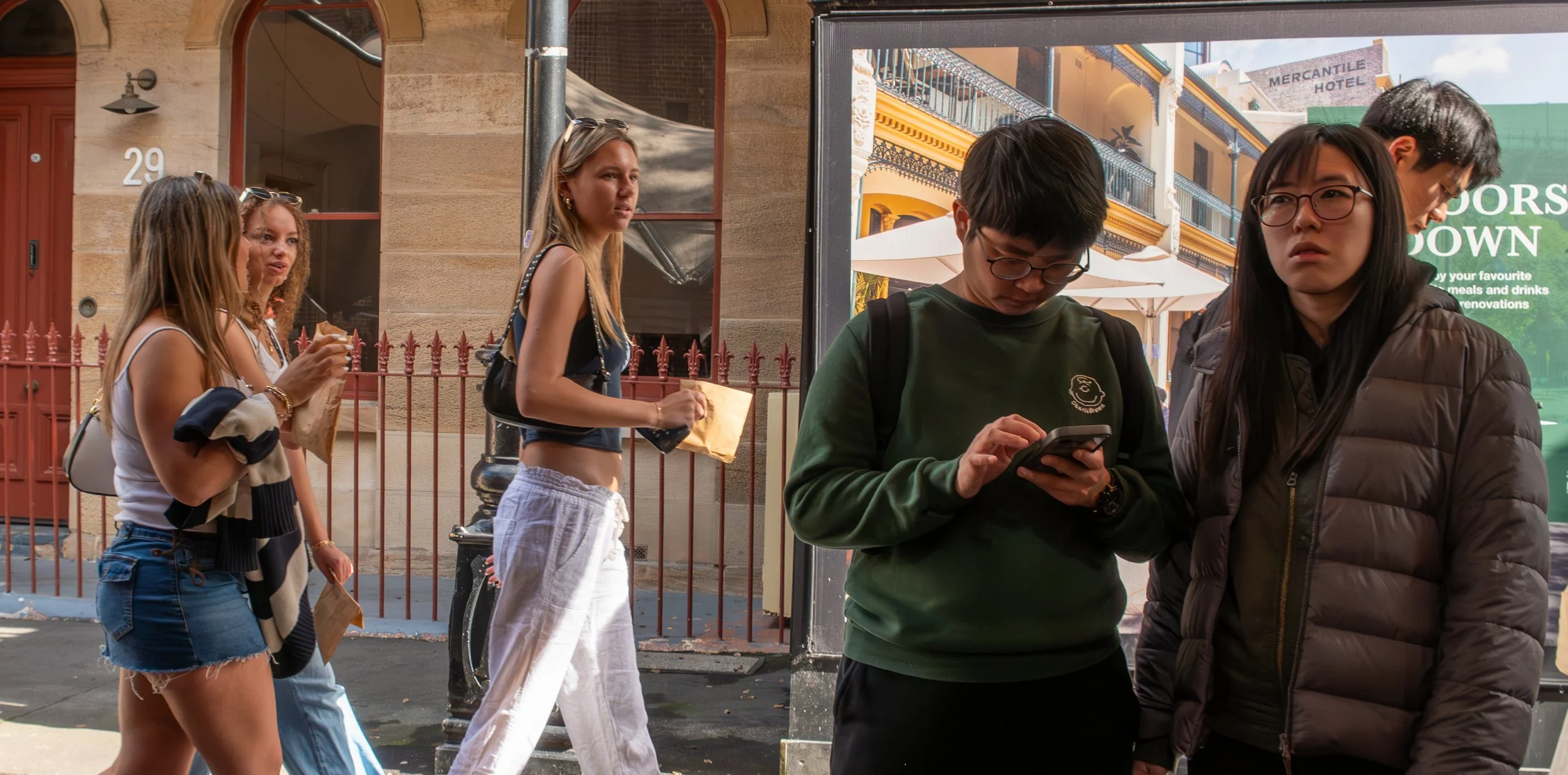 Group of five diverse young women and men standing on a city sidewalk, some holding brown paper bags, with one person looking at a smartphone, and others engaged in conversation, in front of a building with a red door and a street lamp.