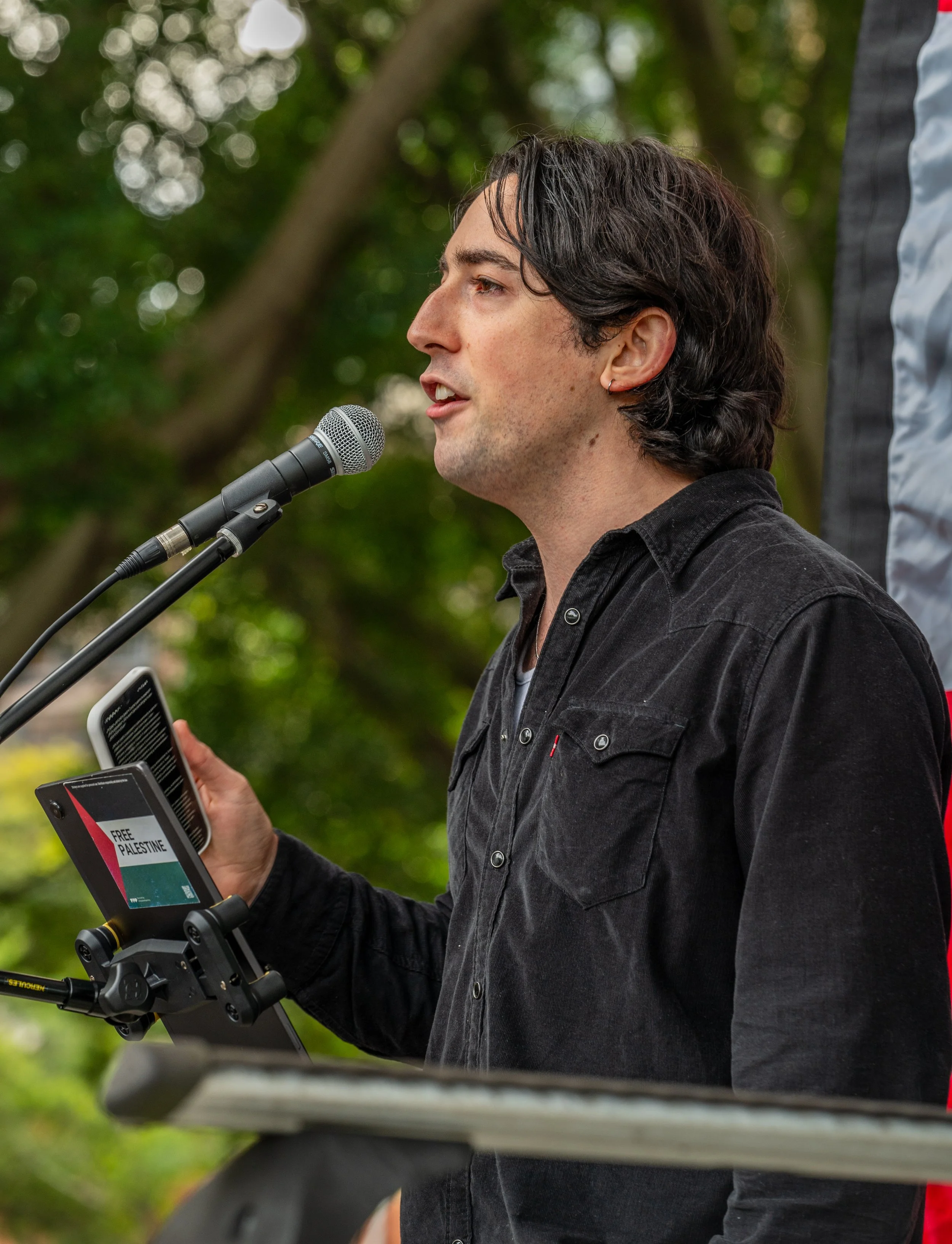 A man with black hair speaking into a microphone outdoors while holding a smartphone showing a flyer for a free Palestine event.