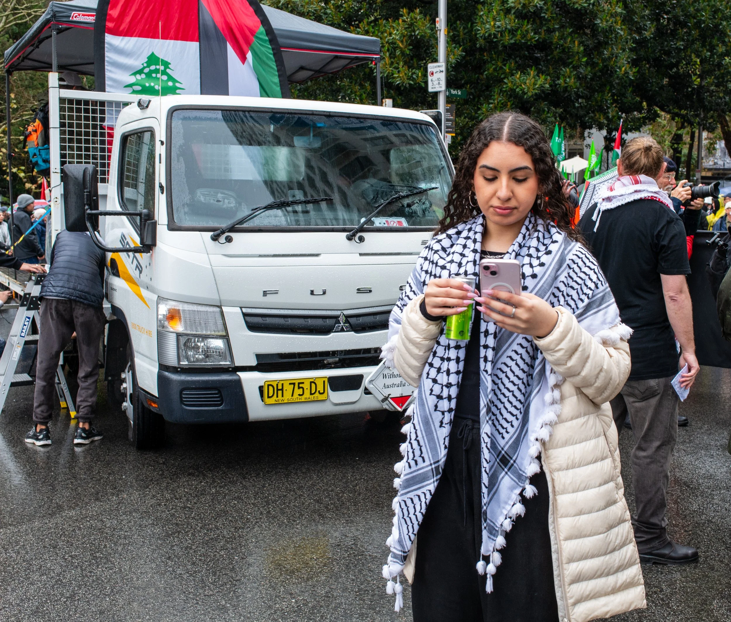A young woman with curly hair, wearing a scarf and a light-colored puffer jacket, is looking at her phone while standing on a wet street at a protest or rally. Behind her, there is a white truck and several other people, some taking photos. The scene appears to be outdoors in an urban area, with trees and protest signs visible.